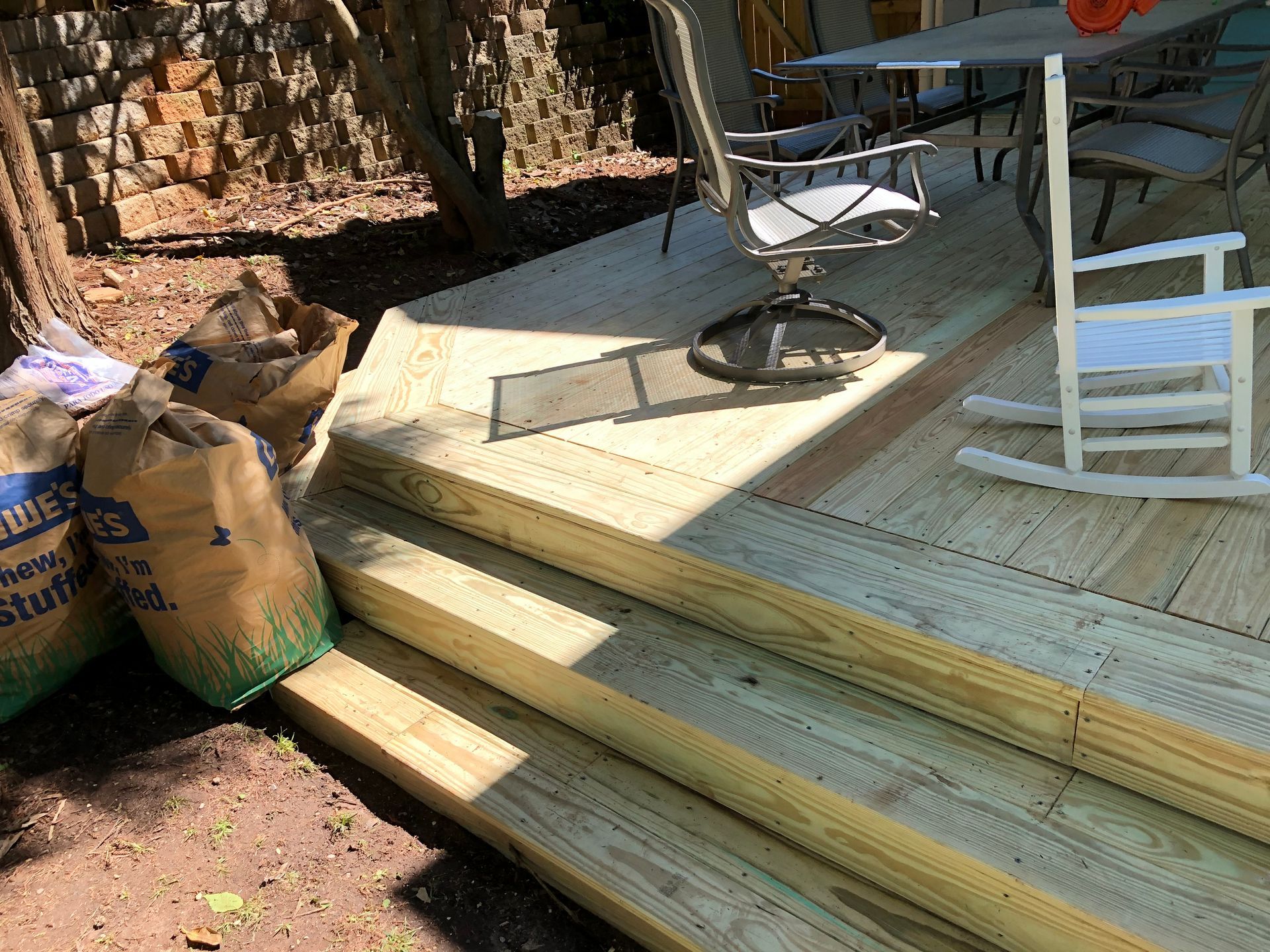 Wooden deck steps leading to a patio with chairs, a table, and bags of soil nearby