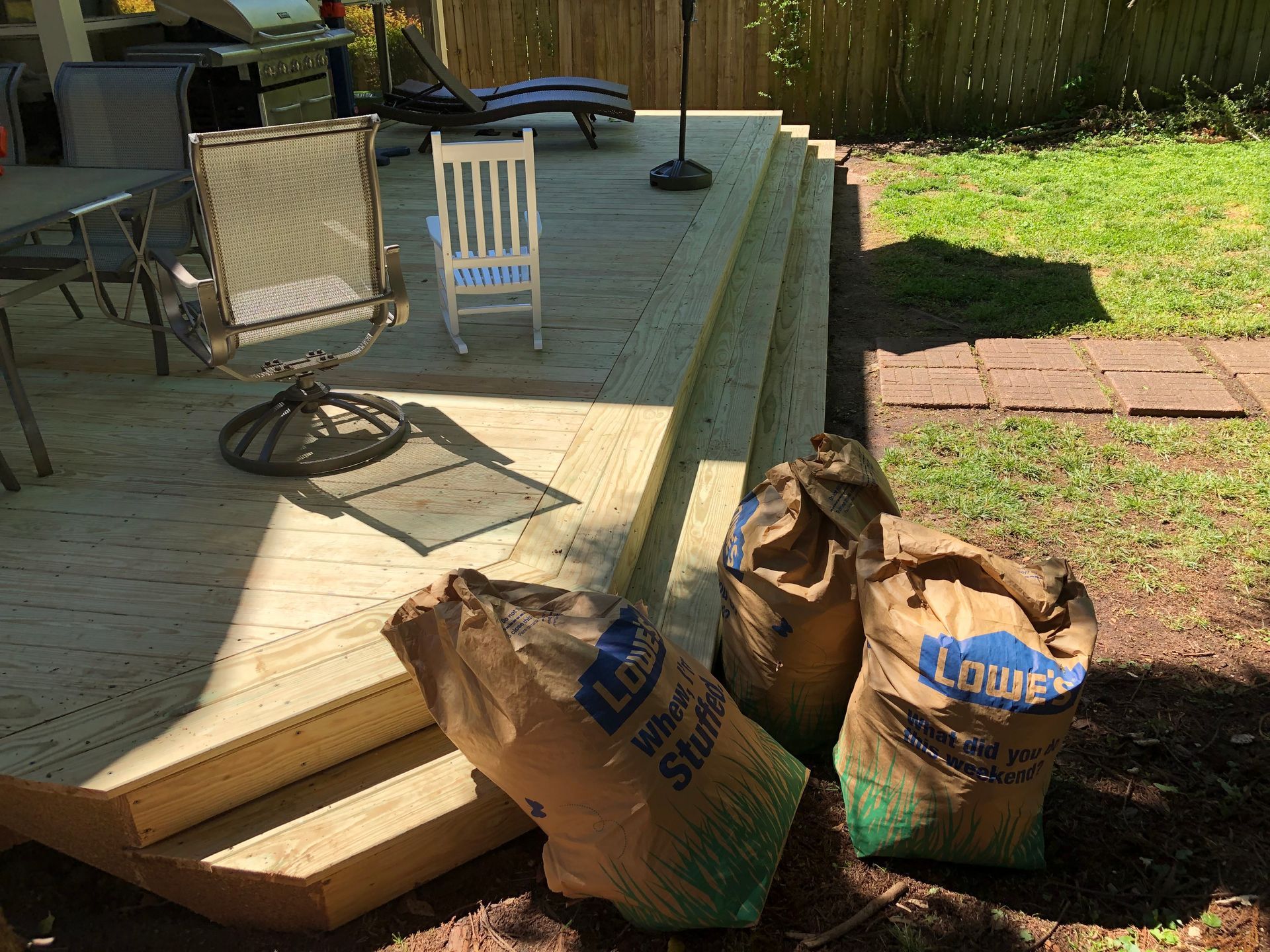 Patio deck with chairs, a lamp, and three mulch bags beside steps leading to a grassy yard