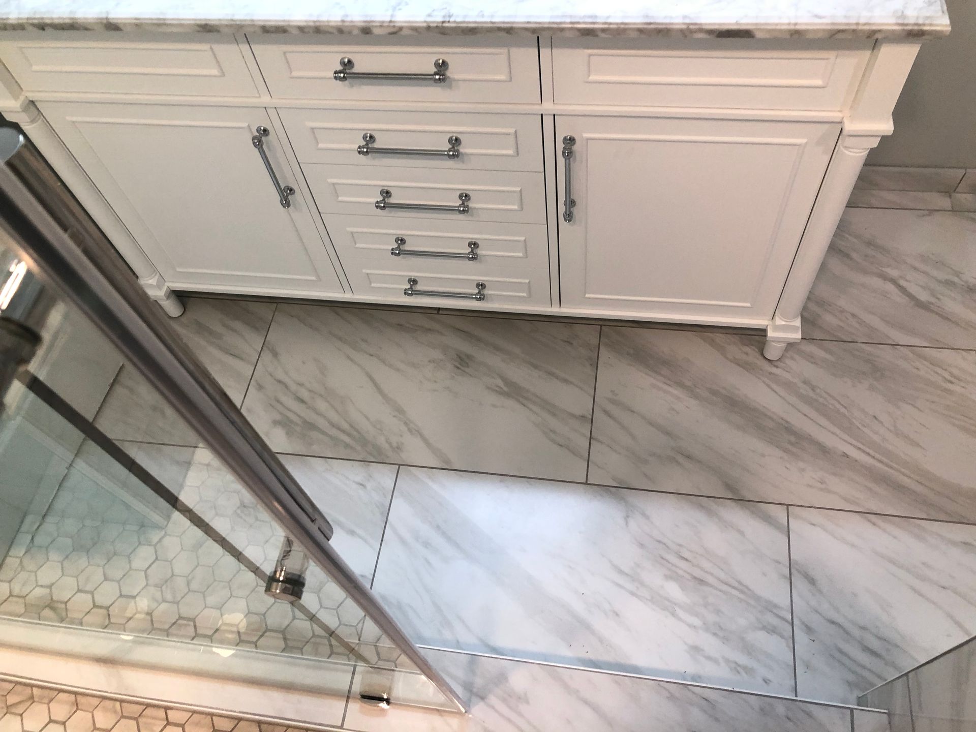 White bathroom vanity with drawers and cabinet beside a glass shower and gray marble tile floor