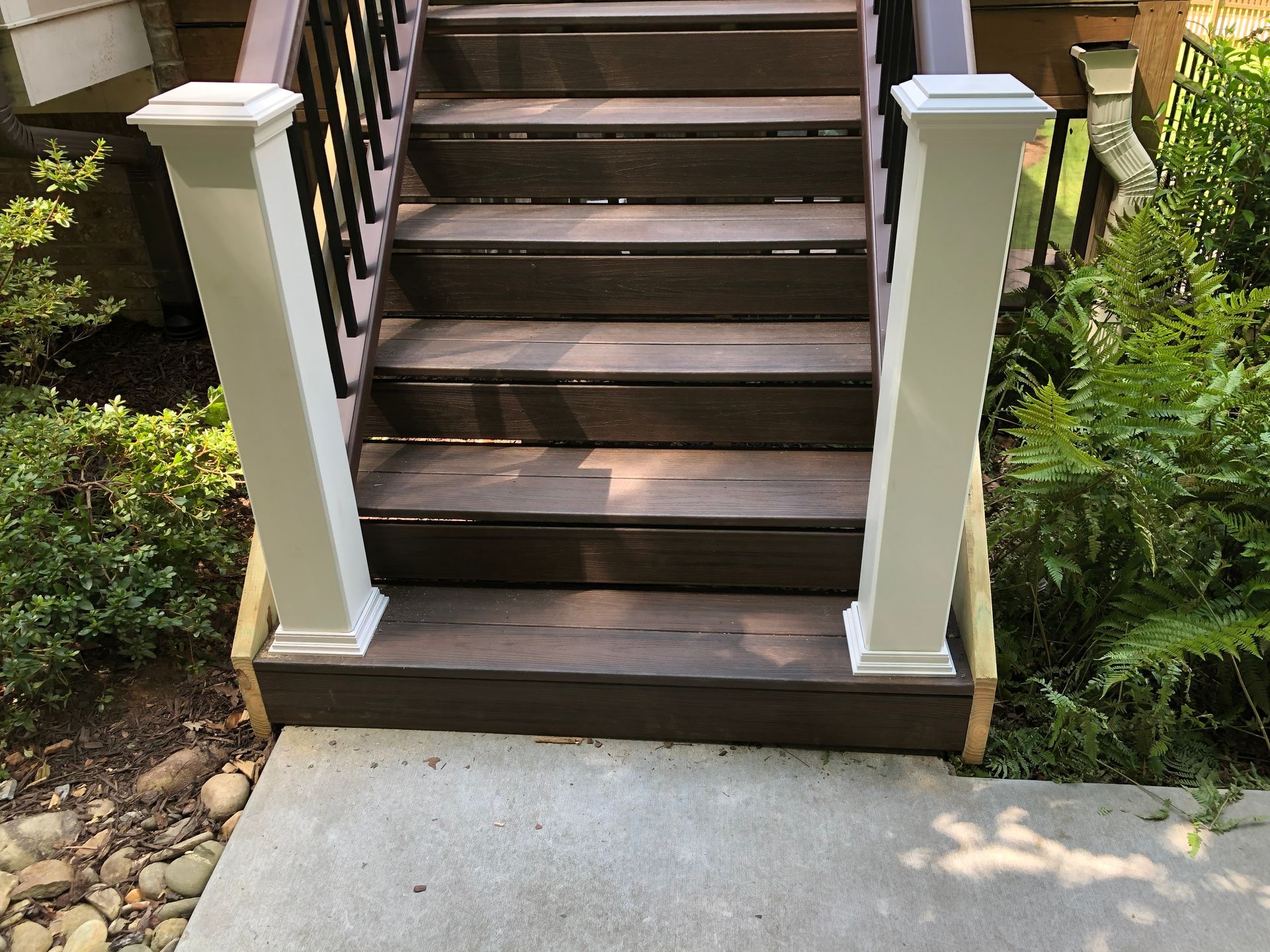 Wooden porch steps with white posts leading up from a concrete walkway, bordered by greenery