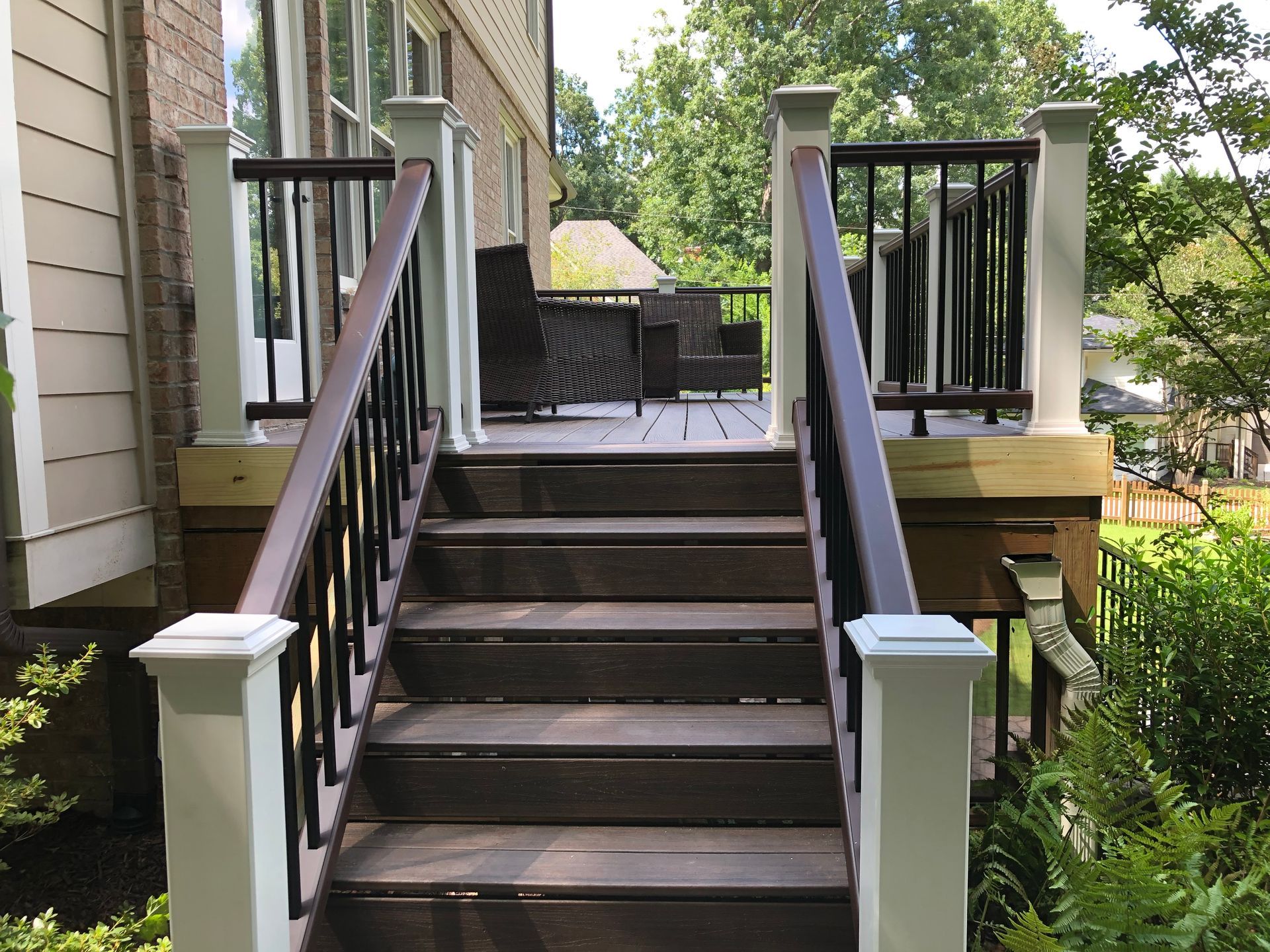 Wooden outdoor stairs with black railings lead to a porch beside a yellow house and green garden.