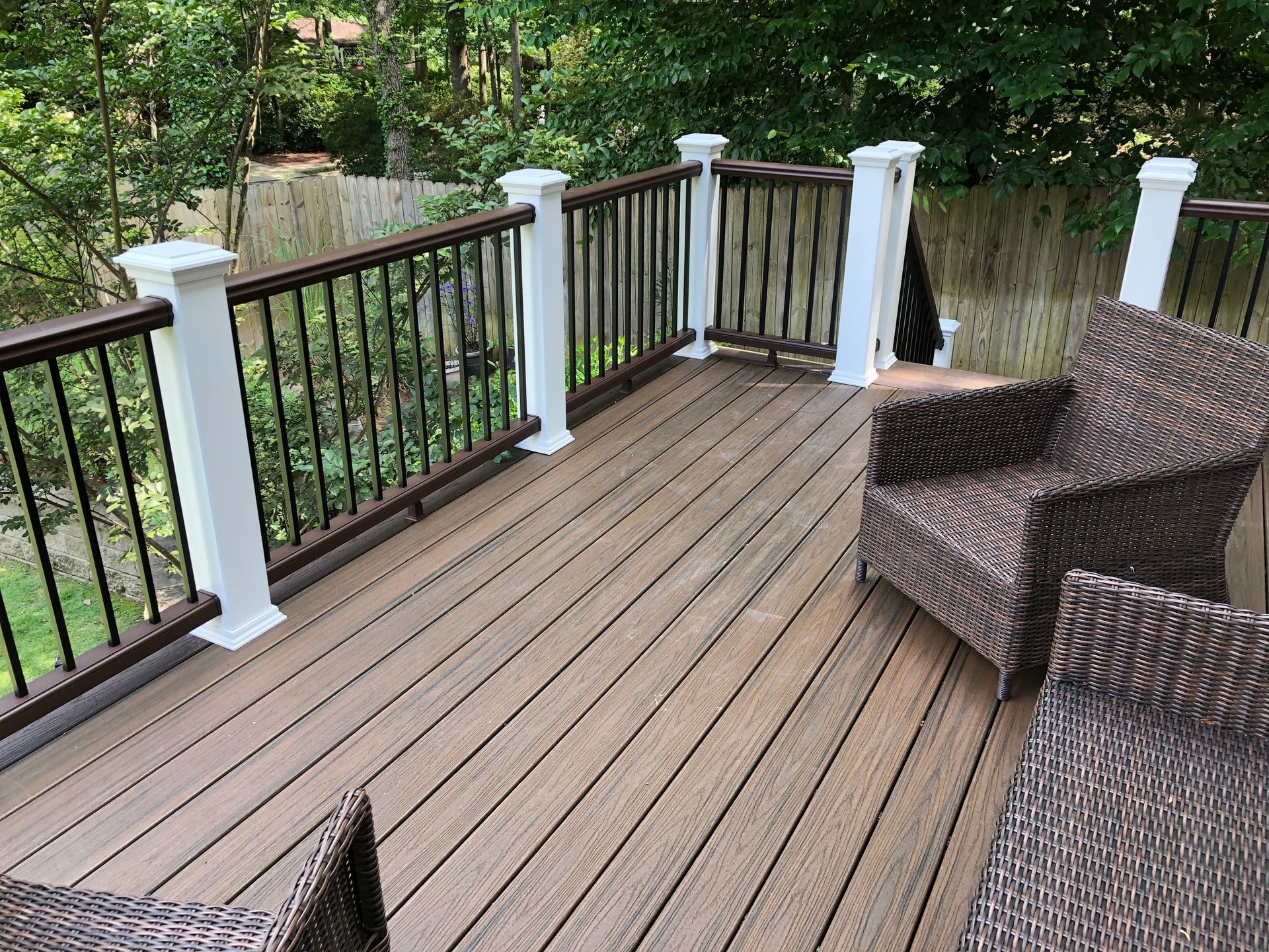 Wooden deck with black railing and white posts, bordered by trees and wicker chairs.