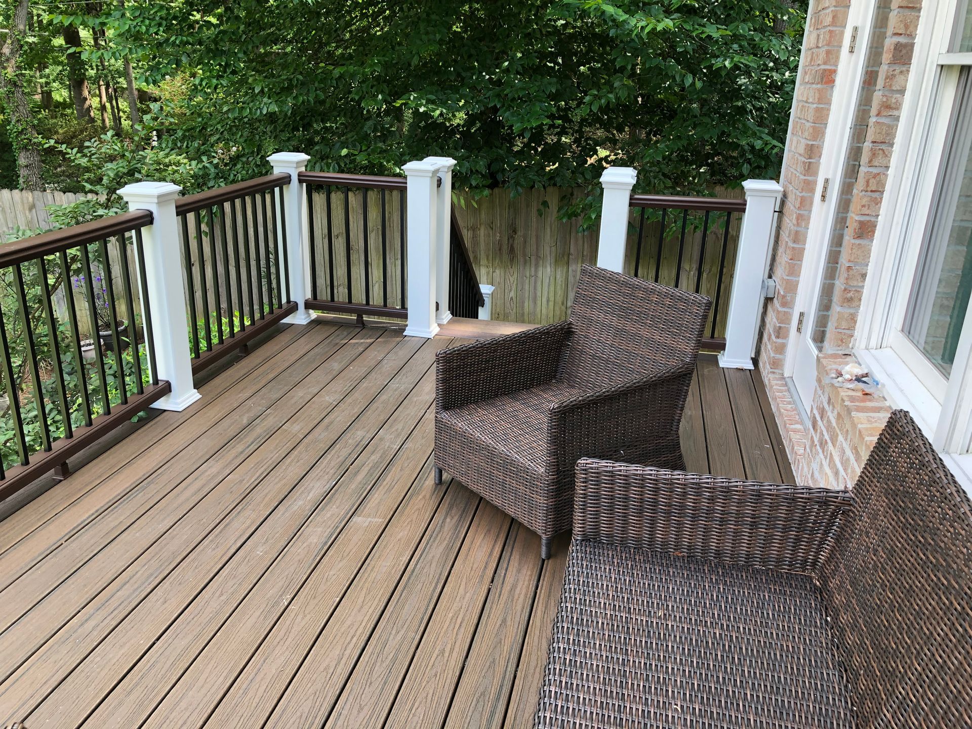 Wooden deck with wicker chairs beside a brick house and white railing, overlooking green trees