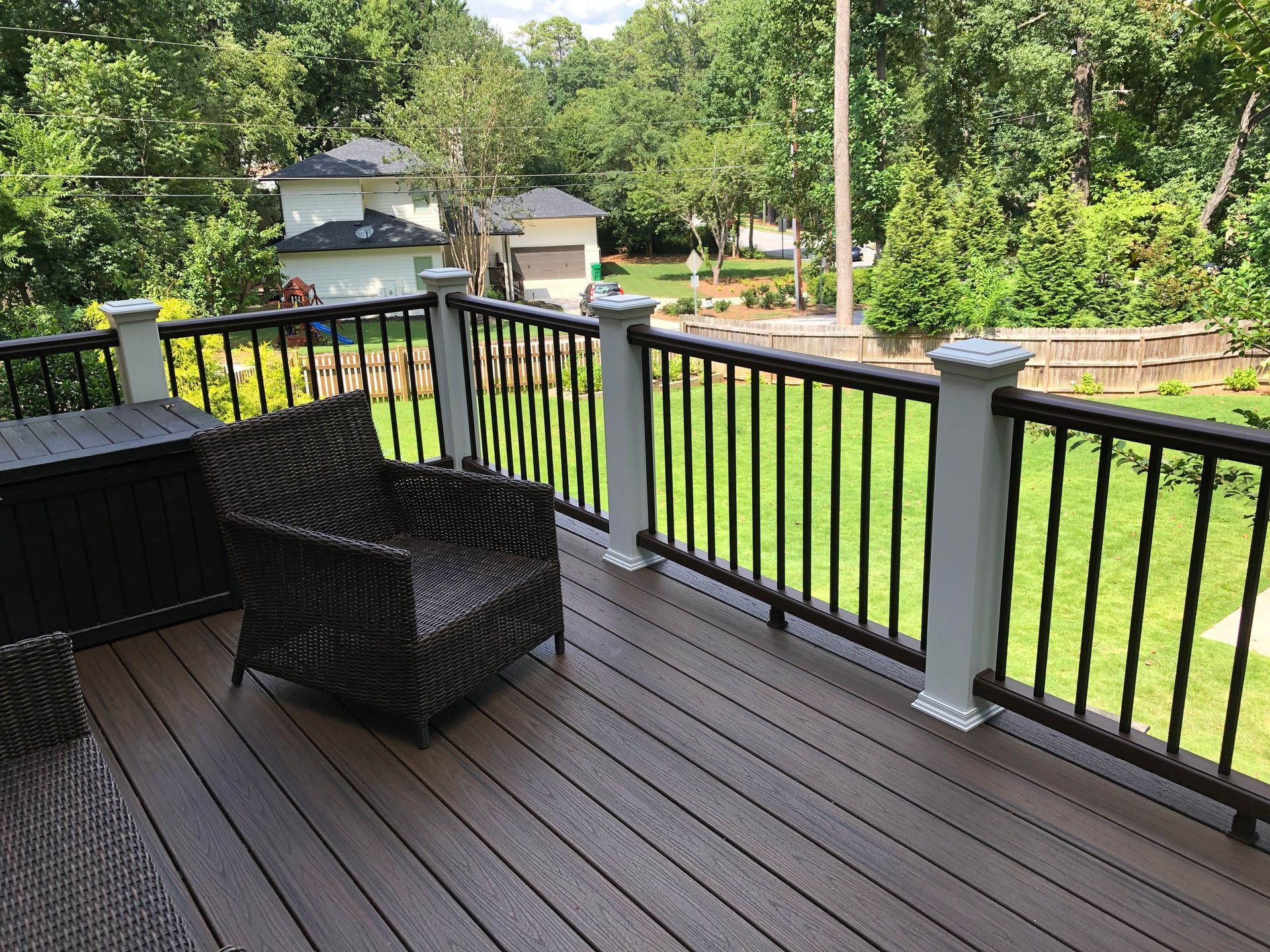 Back deck with black wicker chair, dark railing, and a green yard with trees and houses beyond.