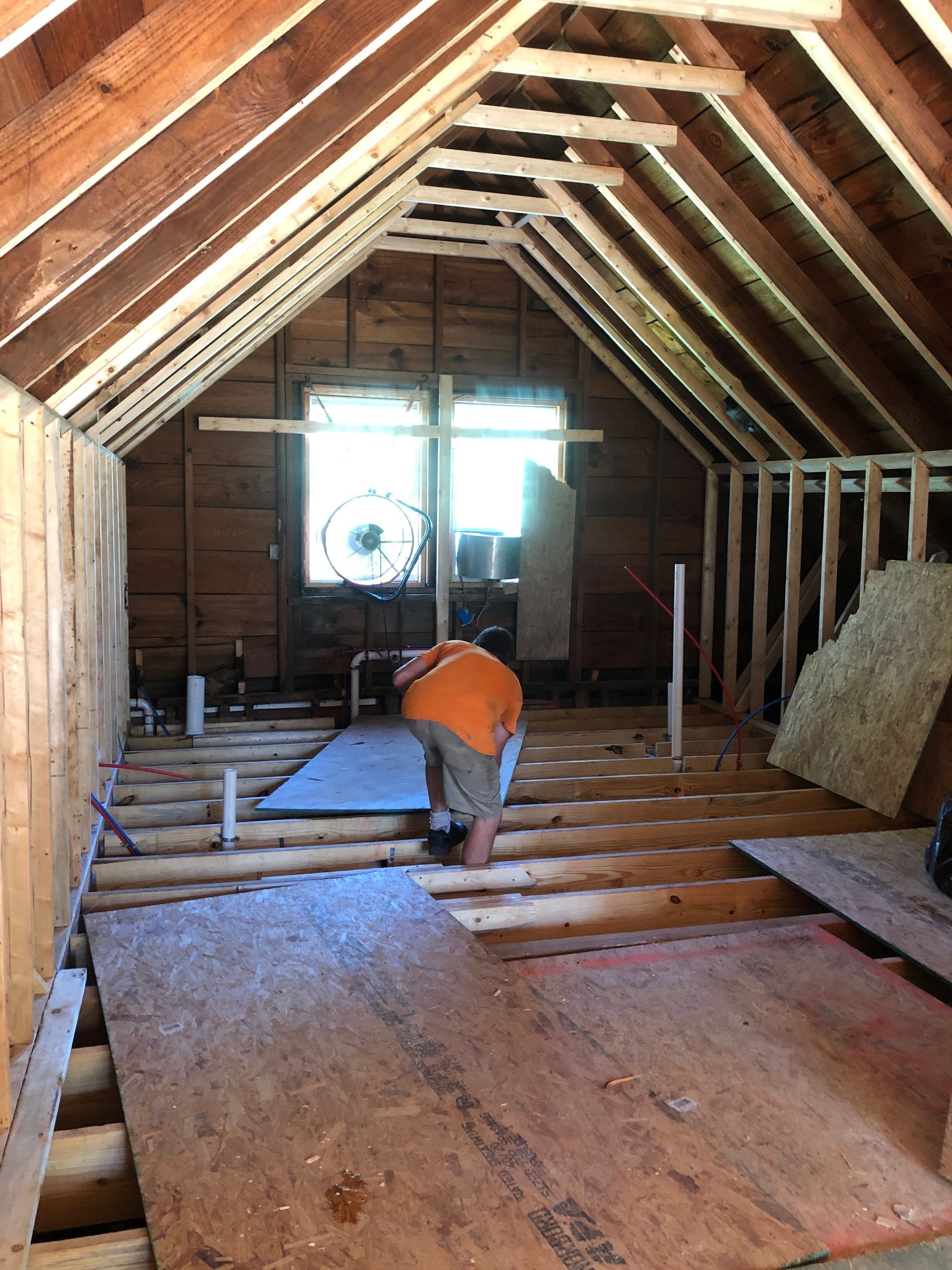 Attic renovation with exposed rafters and a worker in an orange shirt kneeling on floor joists