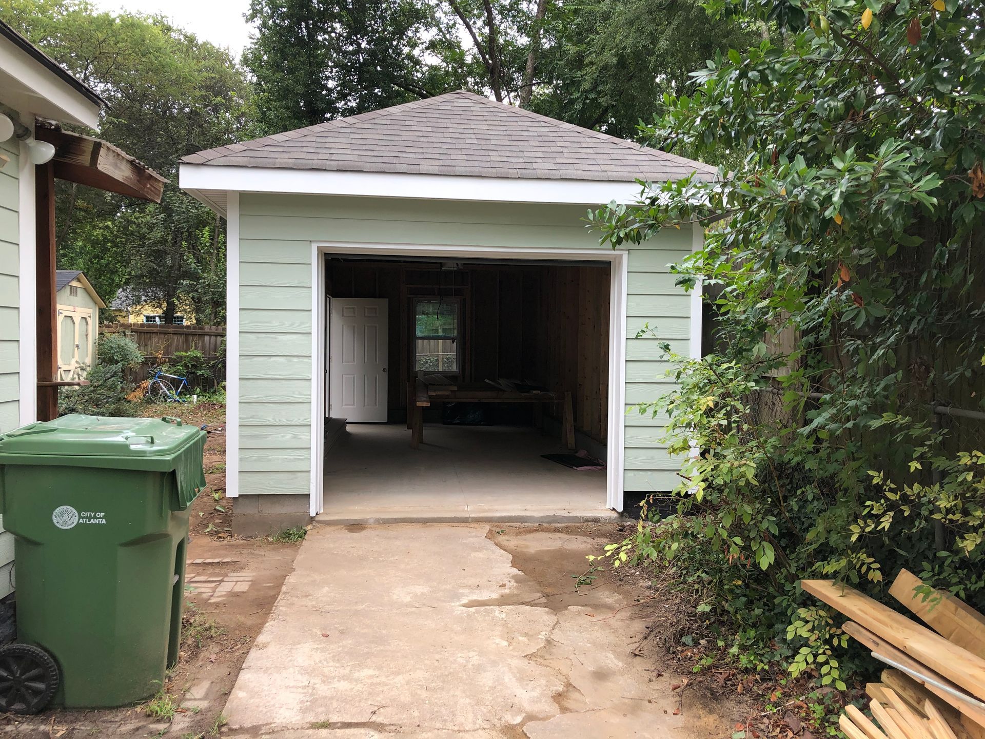 Detached white garage with open door at the end of a narrow concrete driveway, surrounded by trees and bushes