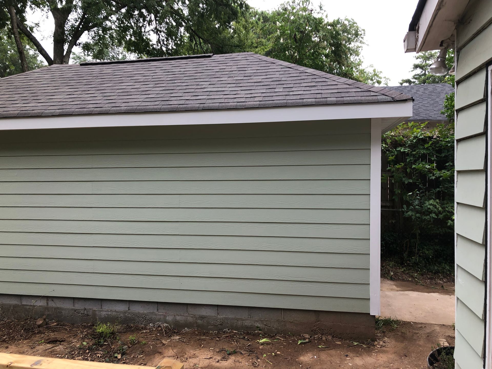 Light gray garage with dark shingled roof beside a narrow walkway and trees.