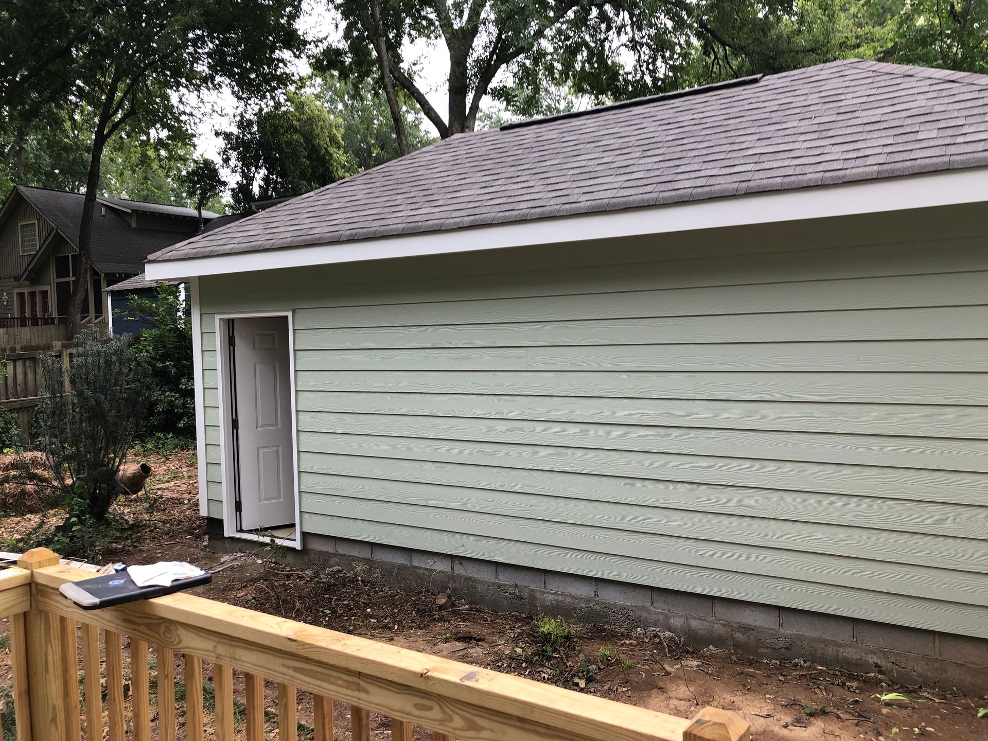 Small white shed with horizontal siding and a gray shingled roof beside a wooden deck and trees.