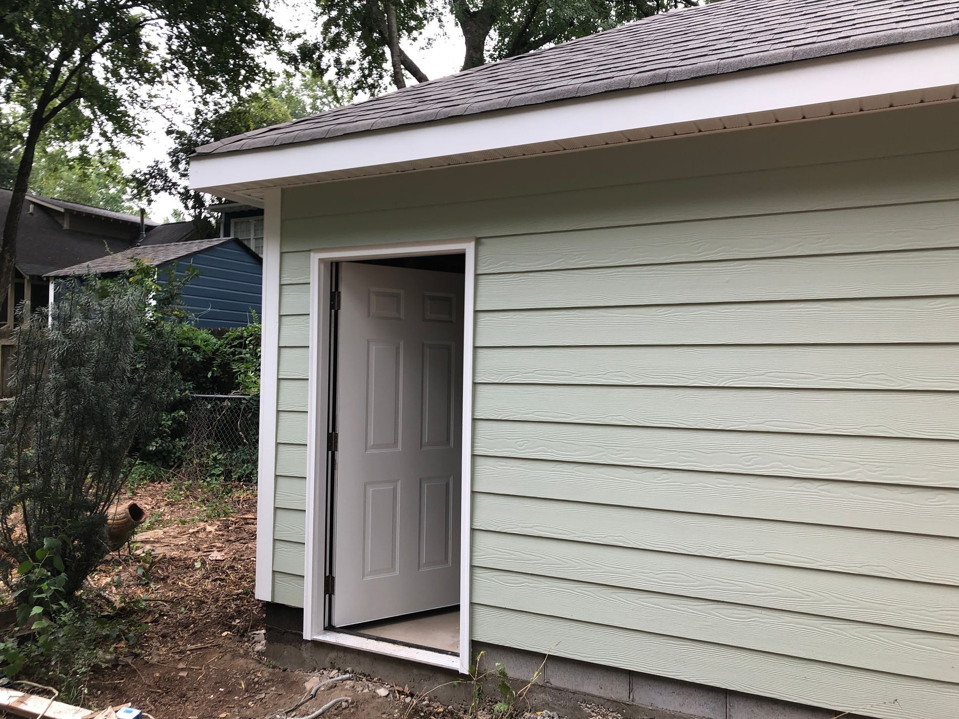 Open exterior door on a white house with horizontal siding and a shingled roof, beside a wooded yard