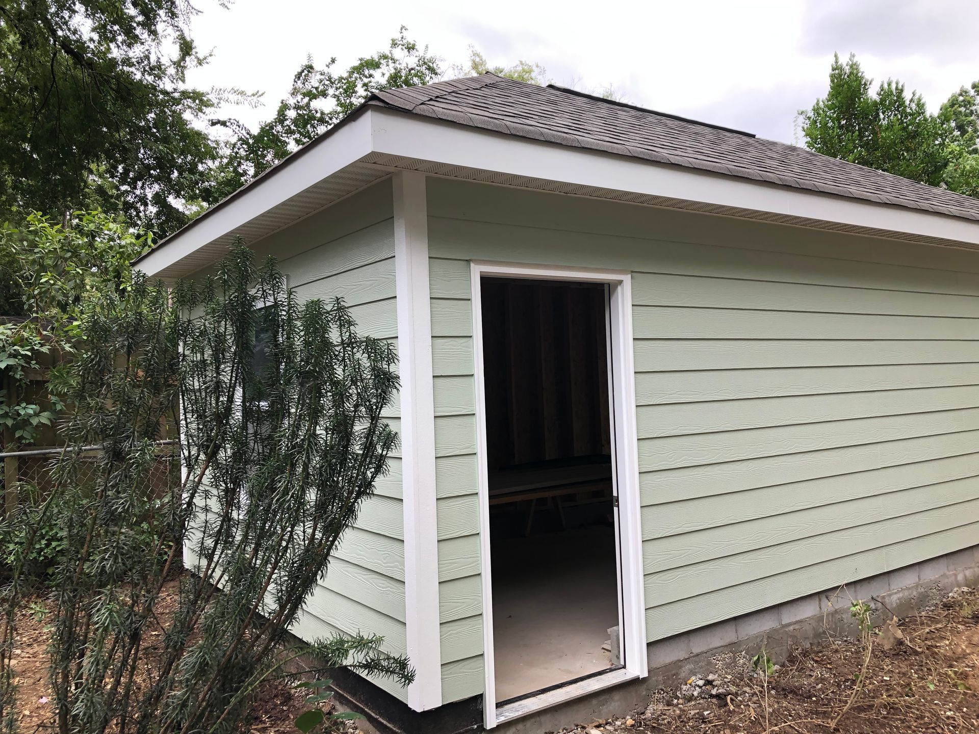 Small light-green shed with white trim and open door in a wooded yard