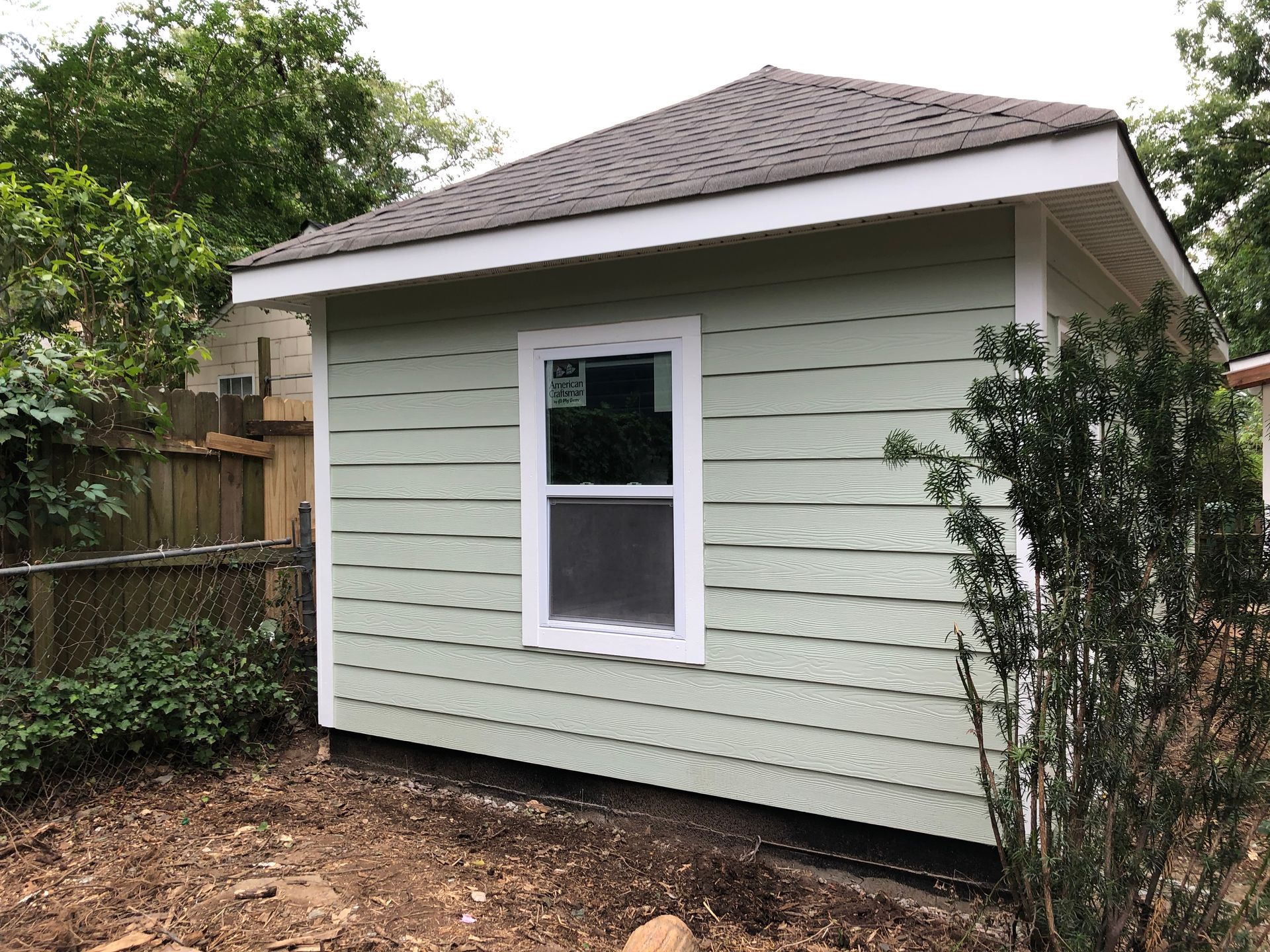 Small light-green shed with a single window and gray shingled roof in a backyard.