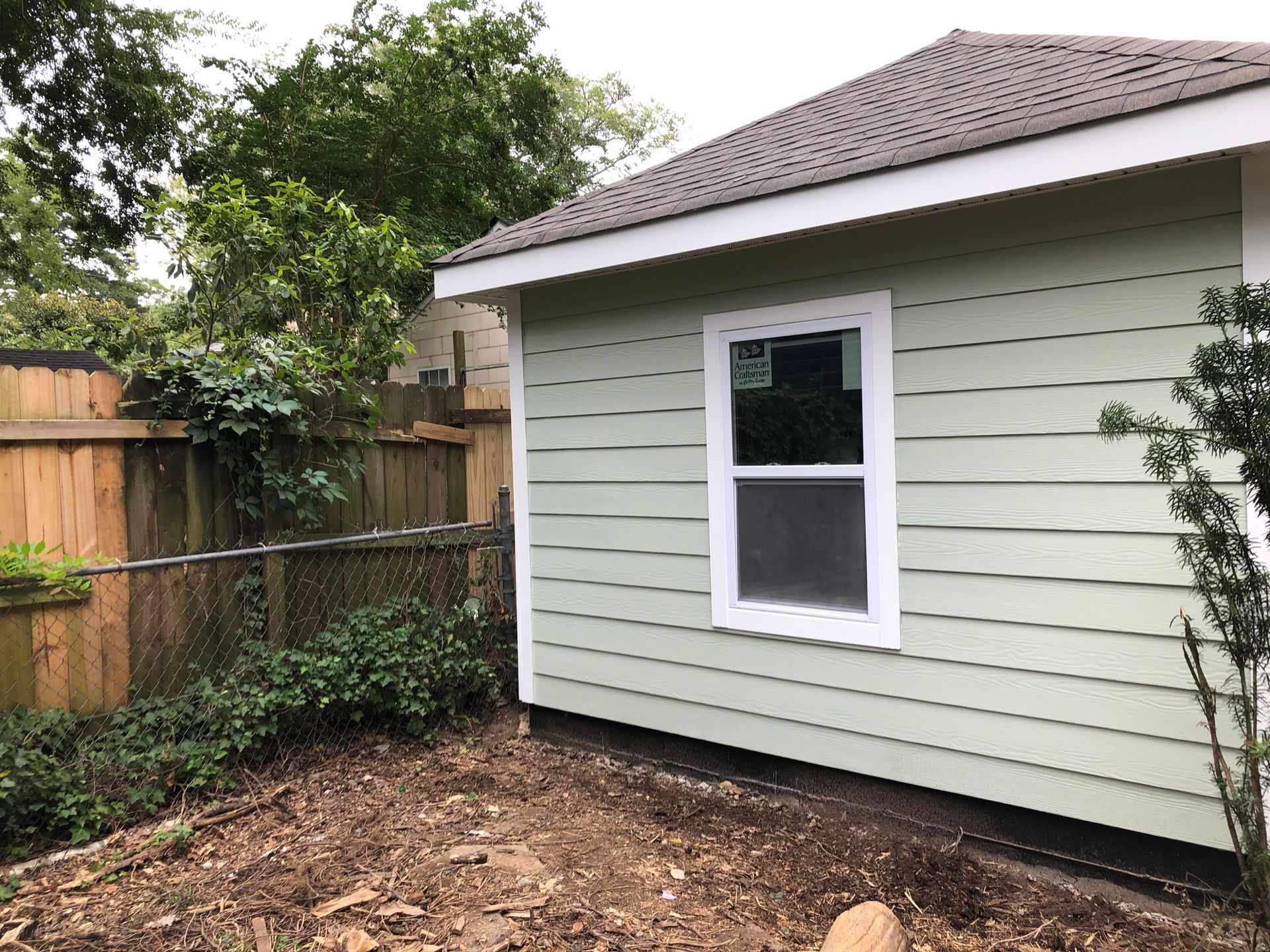 Backyard corner with light green house siding, white window, wooden fence, and dirt ground