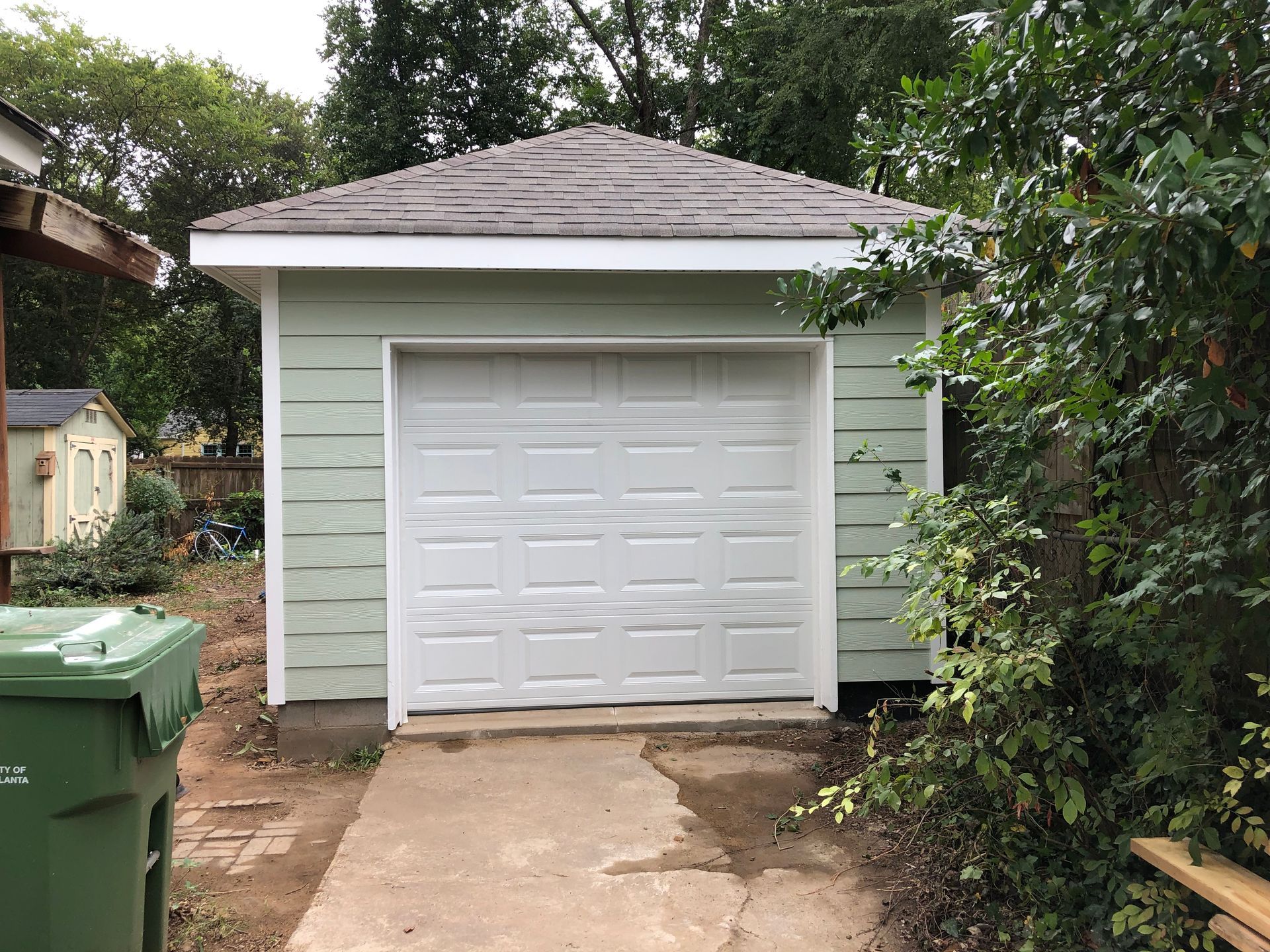 Small green detached garage with white door and driveway, surrounded by trees and bushes