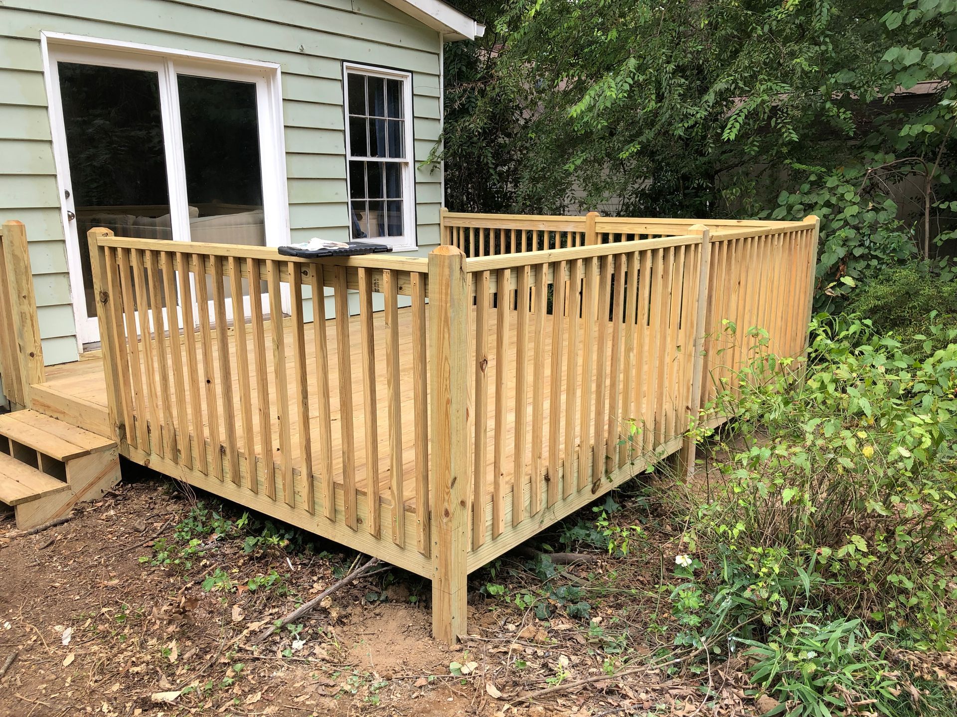 Wooden deck railing under construction beside a house, with unfinished steps and wooded yard.