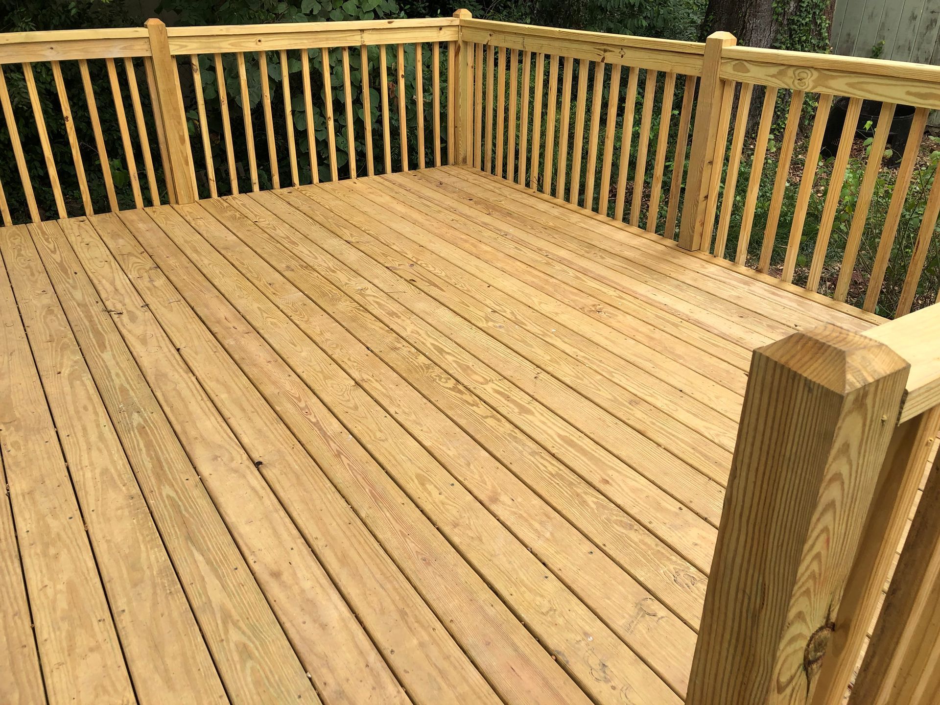 Wooden deck with a tan railing, viewed from one corner in a wooded outdoor setting.