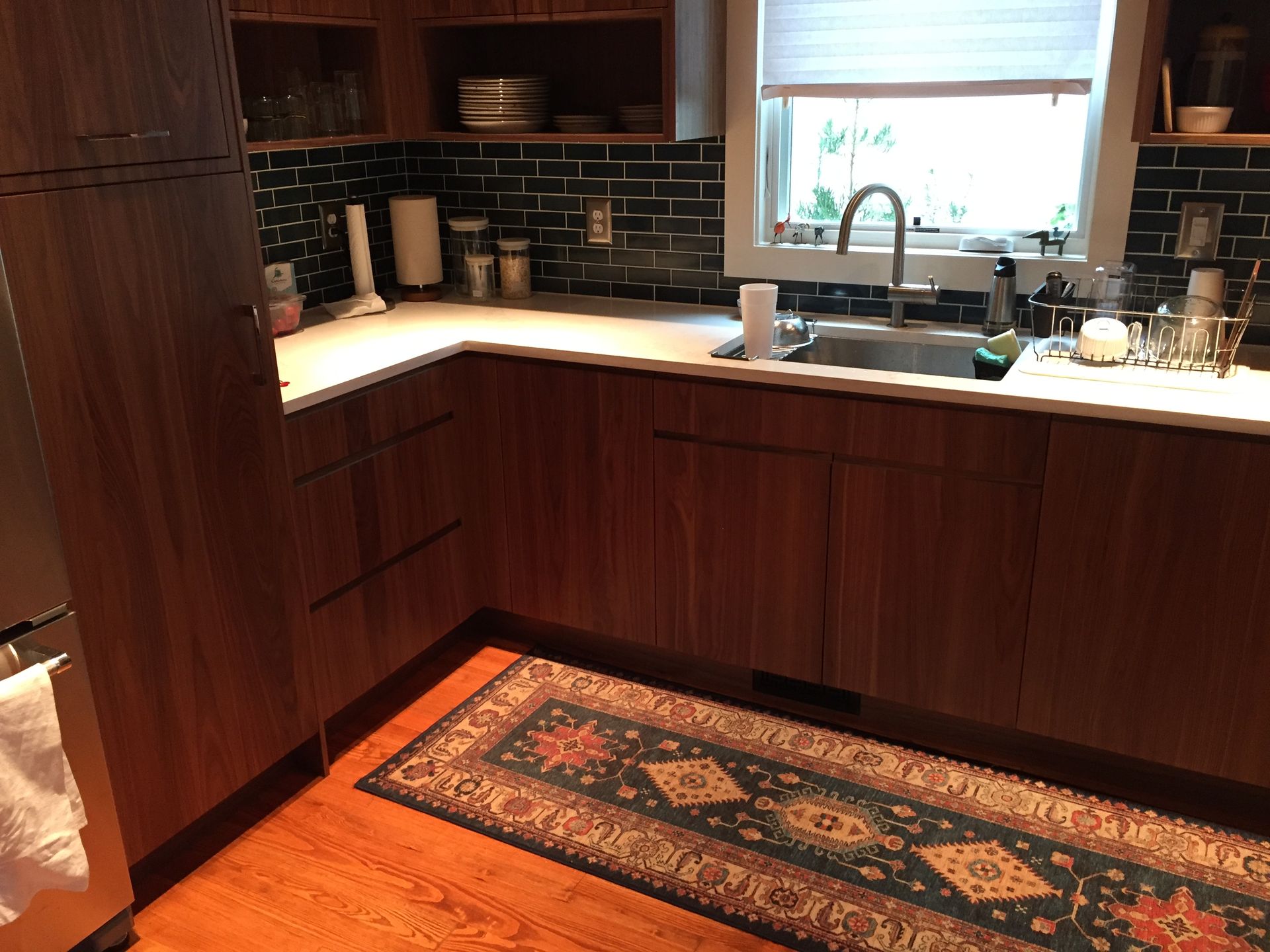 Warm wood kitchen with dark backsplash, white countertop, sink under window, and patterned rug.