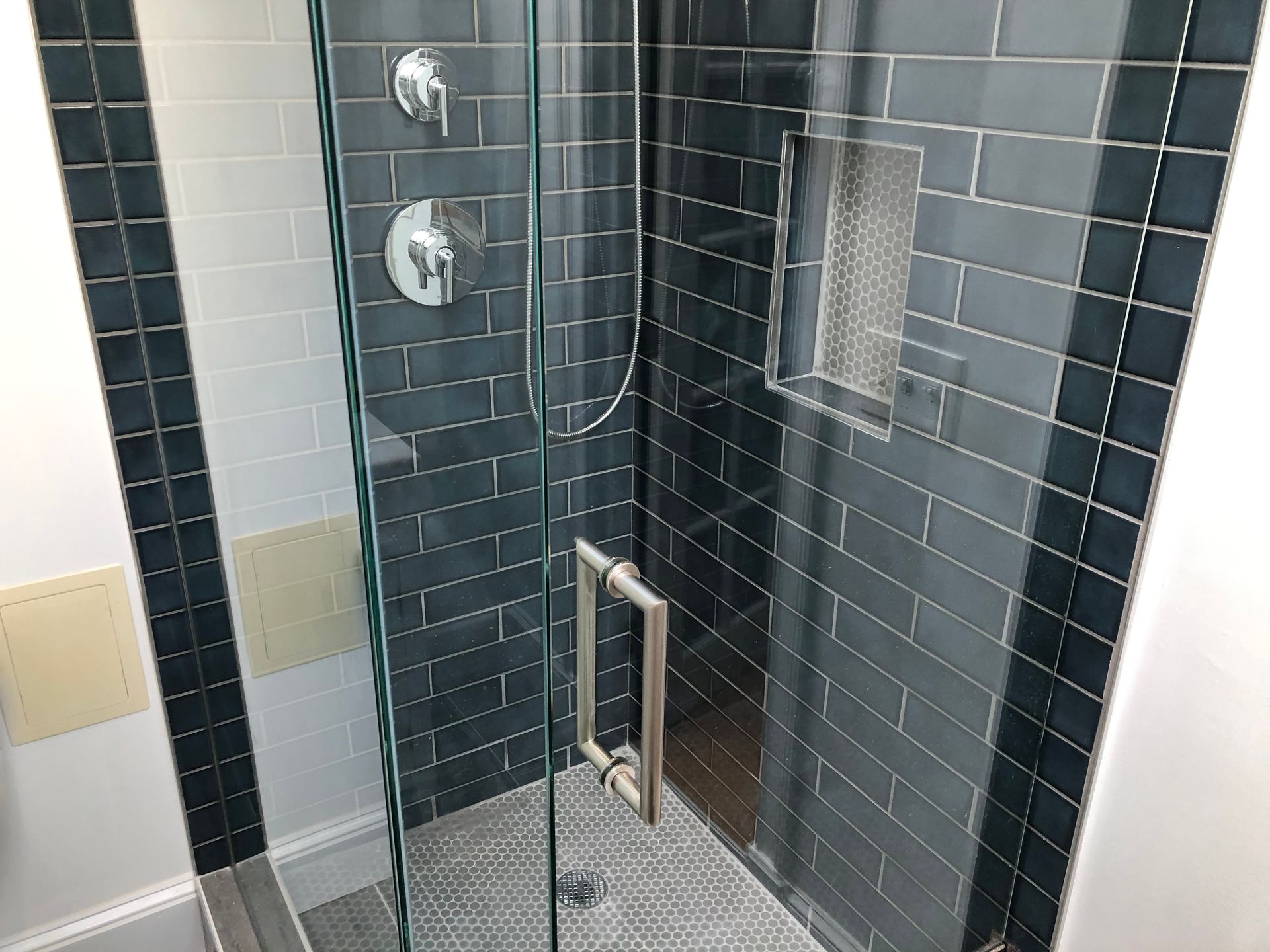Glass shower with dark tile walls, chrome controls, and a small built-in shelf in a tiled bathroom.