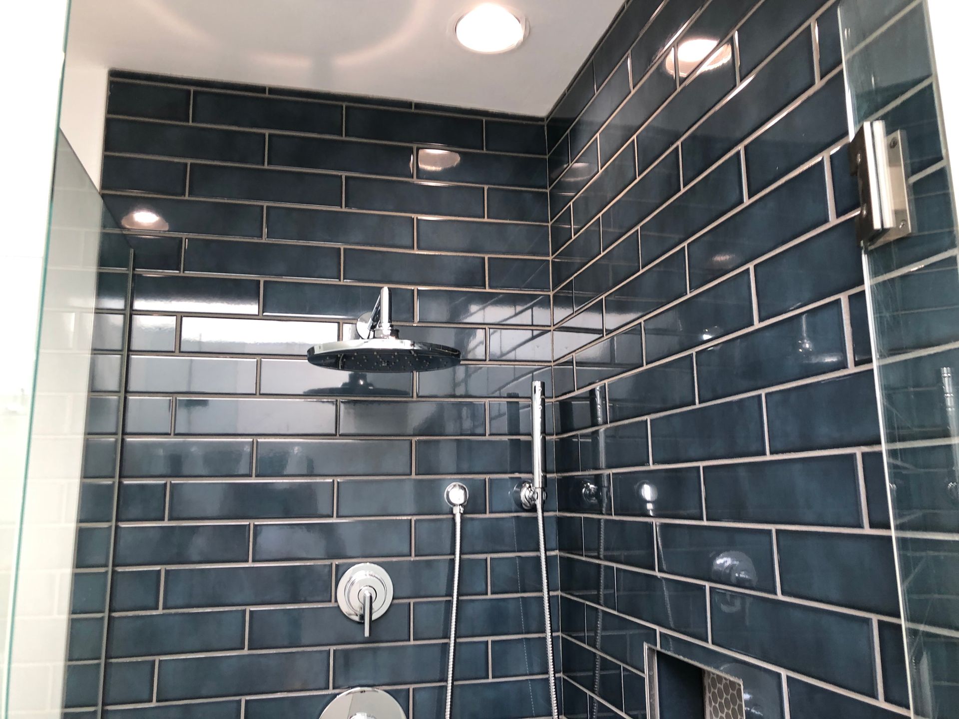 Dark blue tiled shower with glass door, chrome fixtures, and white ceiling light.