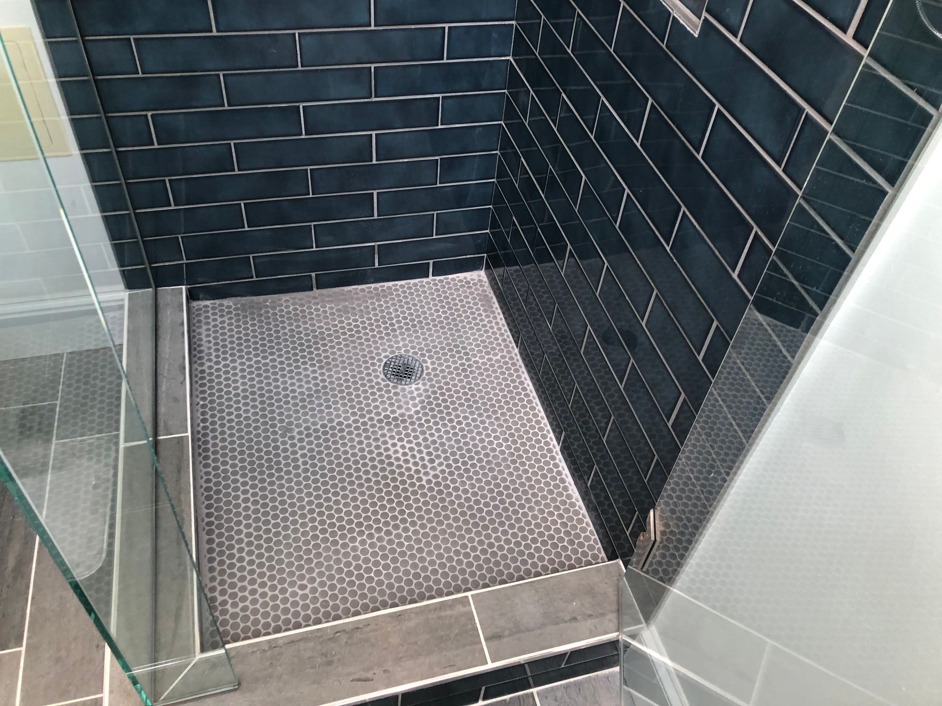 Small tiled shower stall with dark wall tiles, glass door, and a textured gray shower floor mat