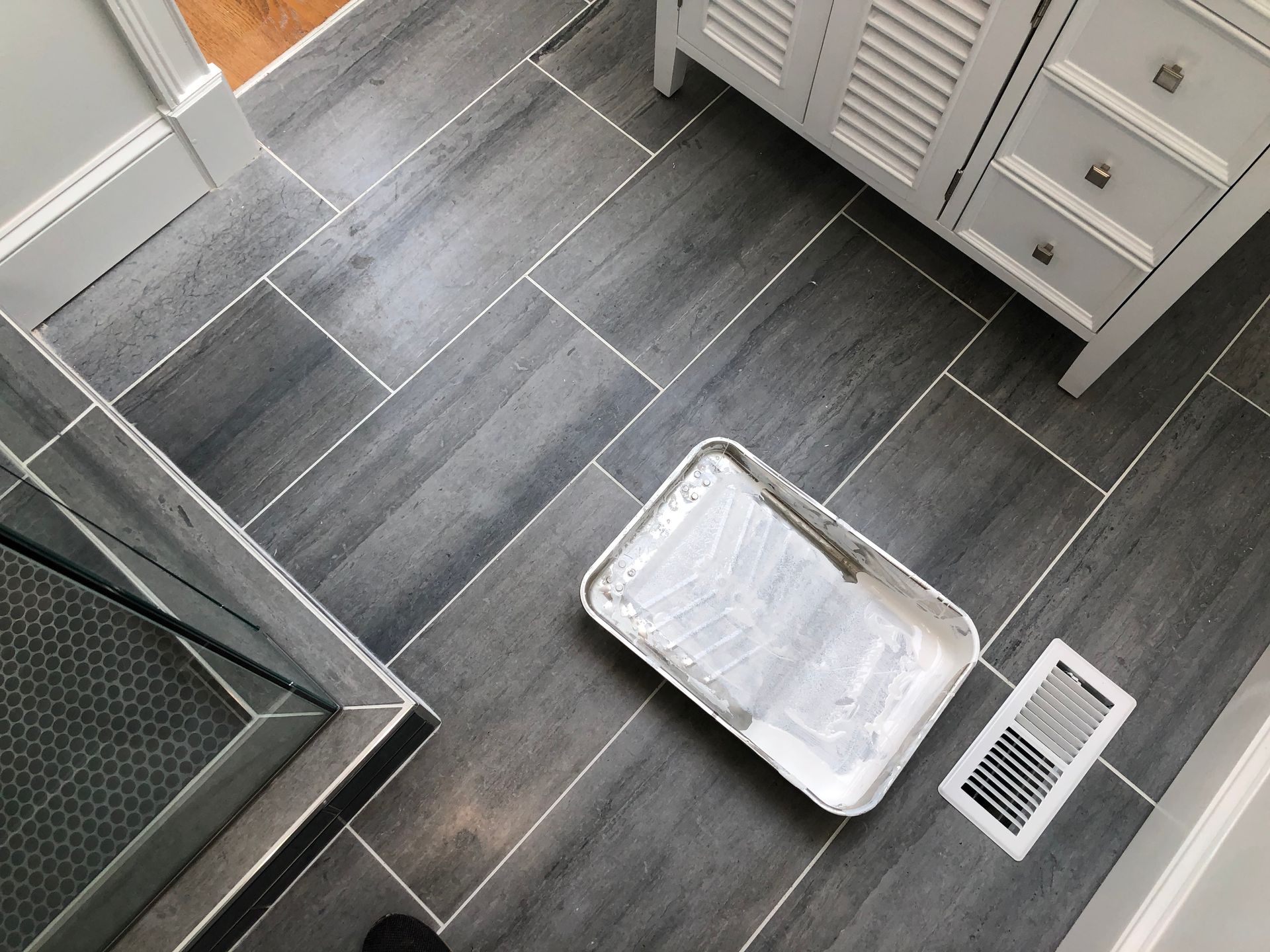 Small bathroom with dark tile floor, shower mat, paint tray, and floor vent.