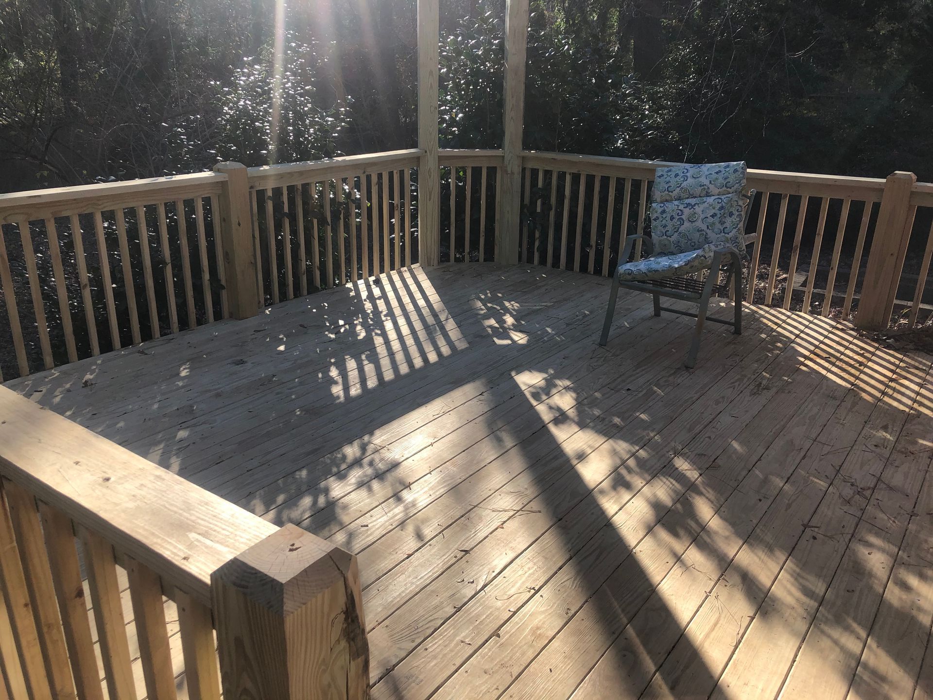 Sunlit wooden deck with railings and a blue chair, casting long shadows through the trees.