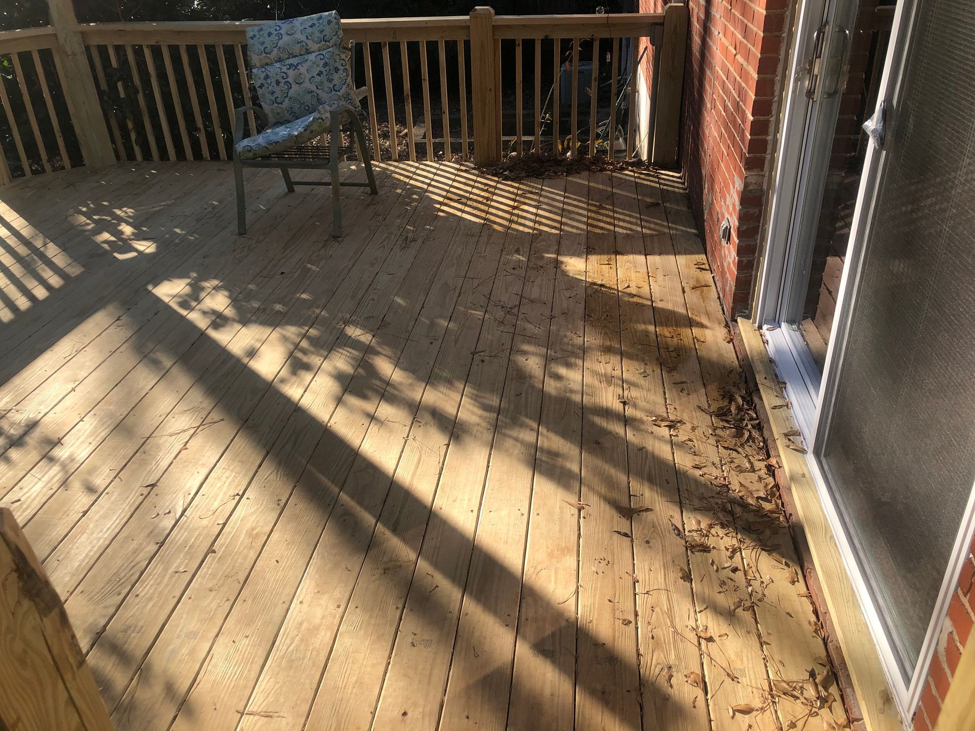 Sunlit wooden deck with railing, a chair, and long diagonal shadows beside a brick wall.