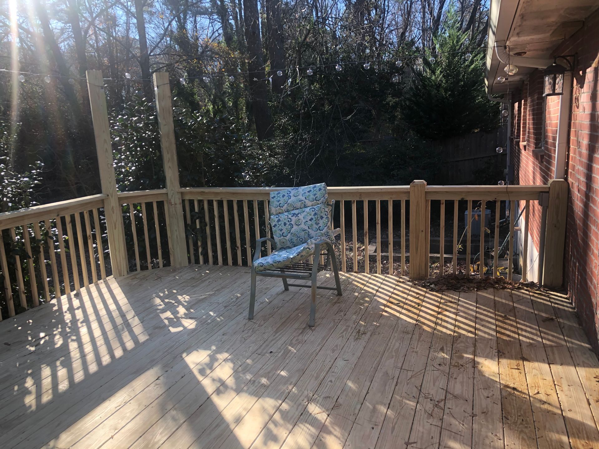 Wooden deck with a single chair in sunlight, overlooking trees and a brick house wall.