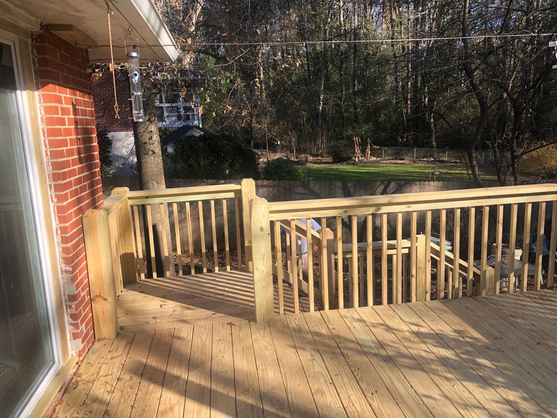Wooden deck with railing beside a brick house, overlooking a wooded backyard in bright sunlight