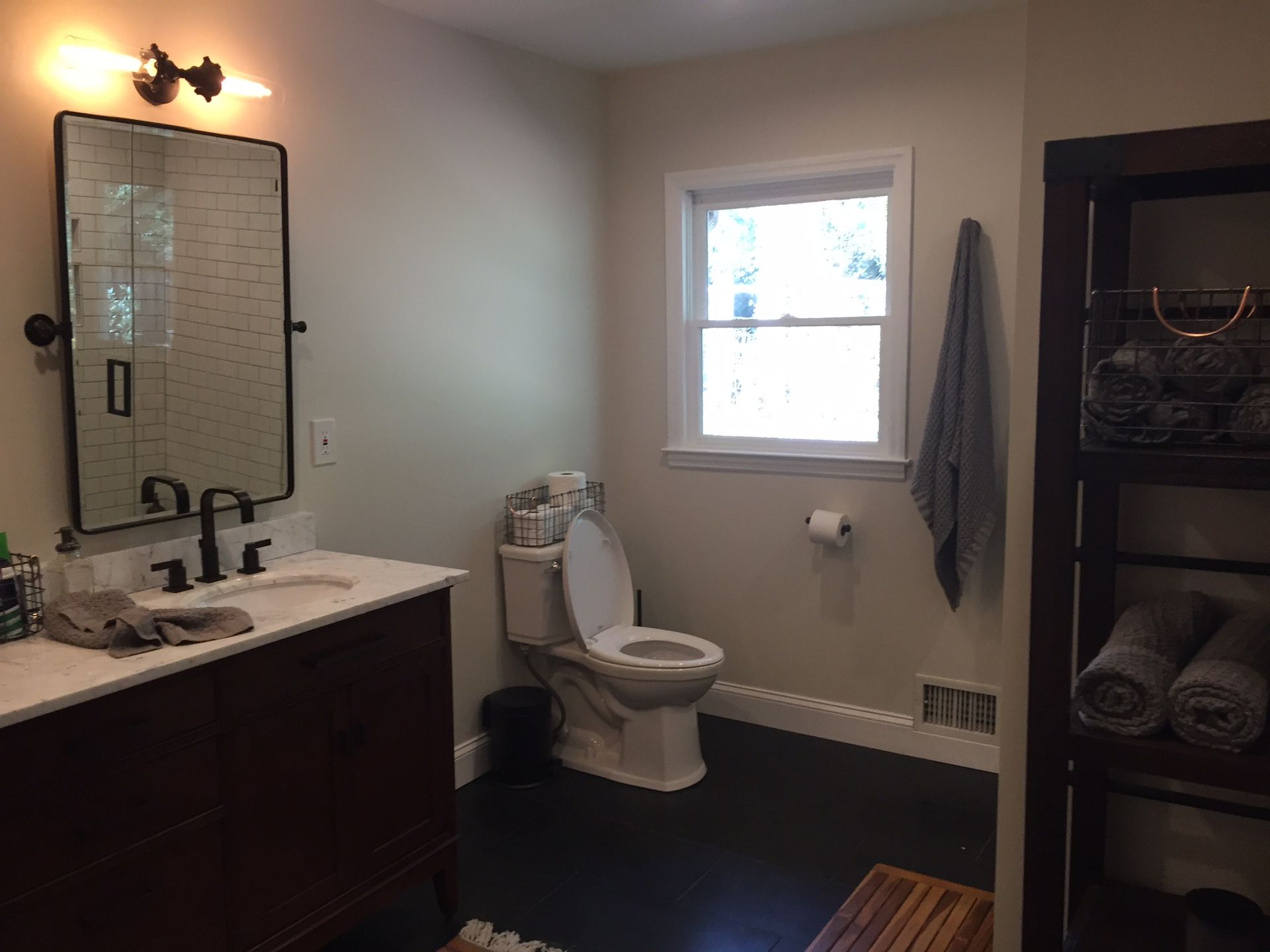 Bathroom with sink and mirror on left, toilet by window, towel rack, and dark shelving on right.