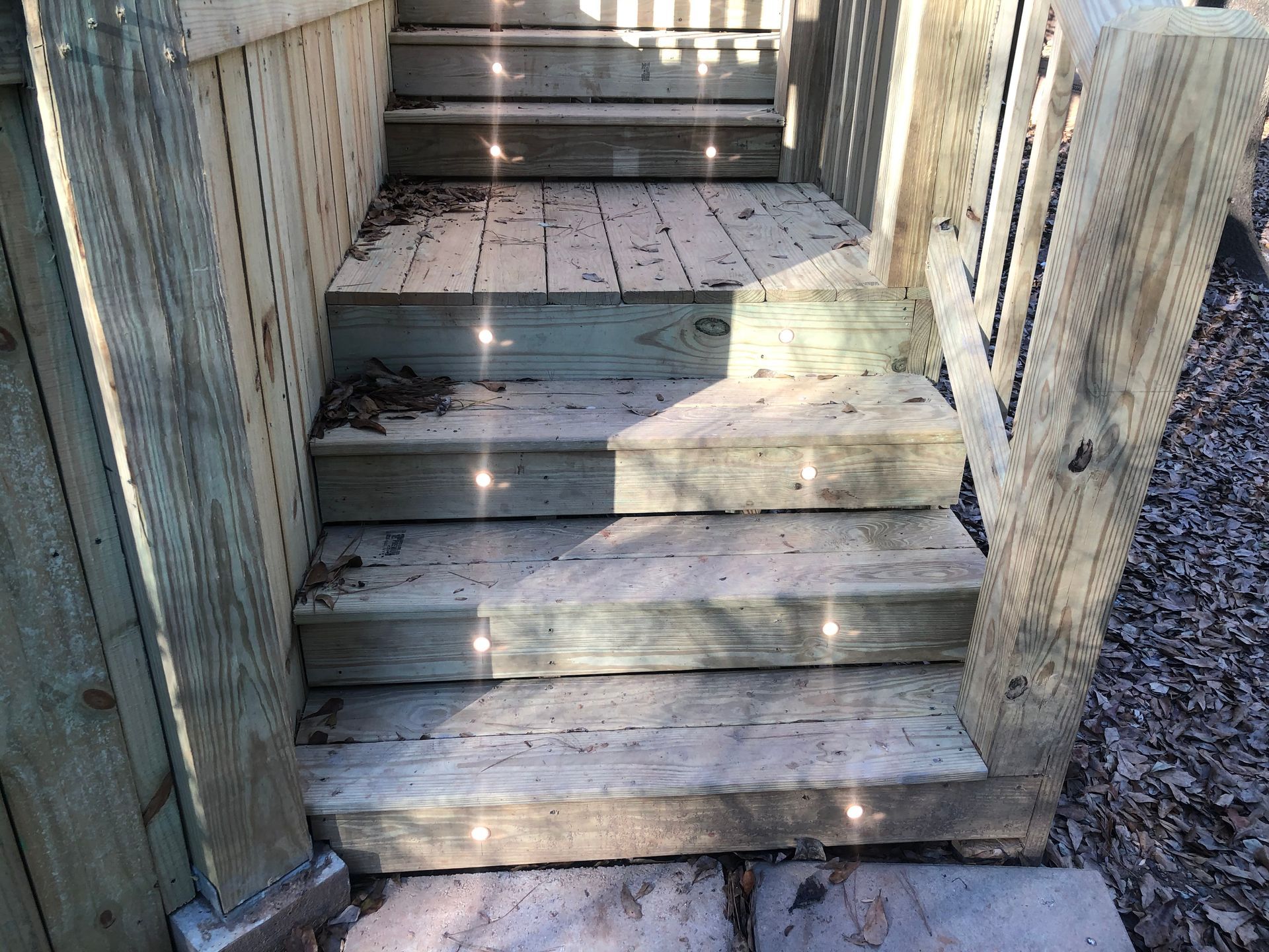 Weathered wooden stairs leading up to a deck, with railings on both sides and sunlight casting shadows.