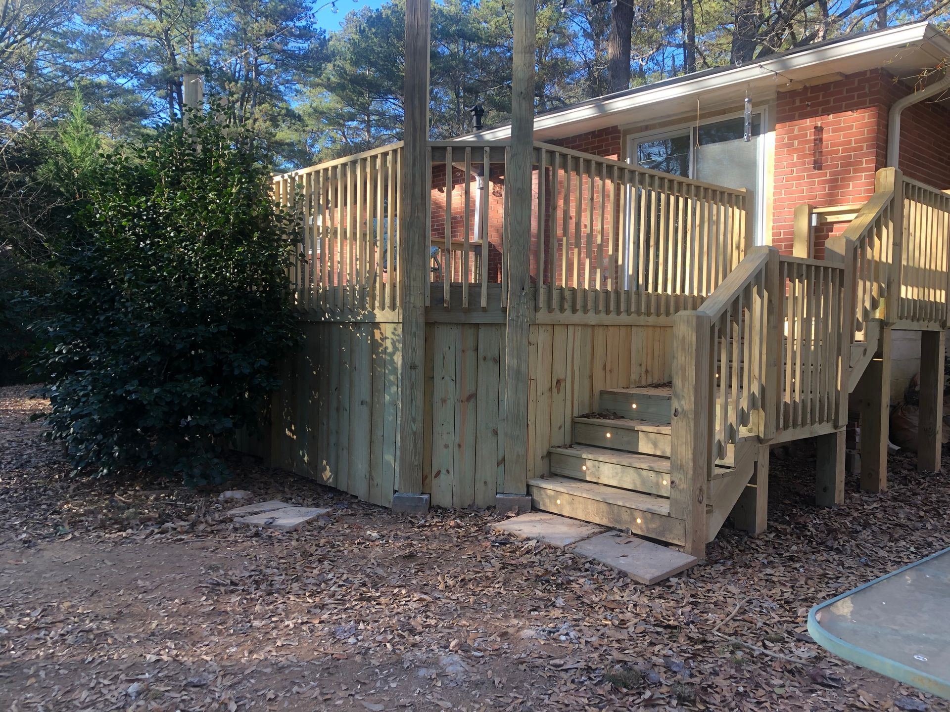 Wooden deck with steps beside a brick house in a wooded yard, with fallen leaves on the ground