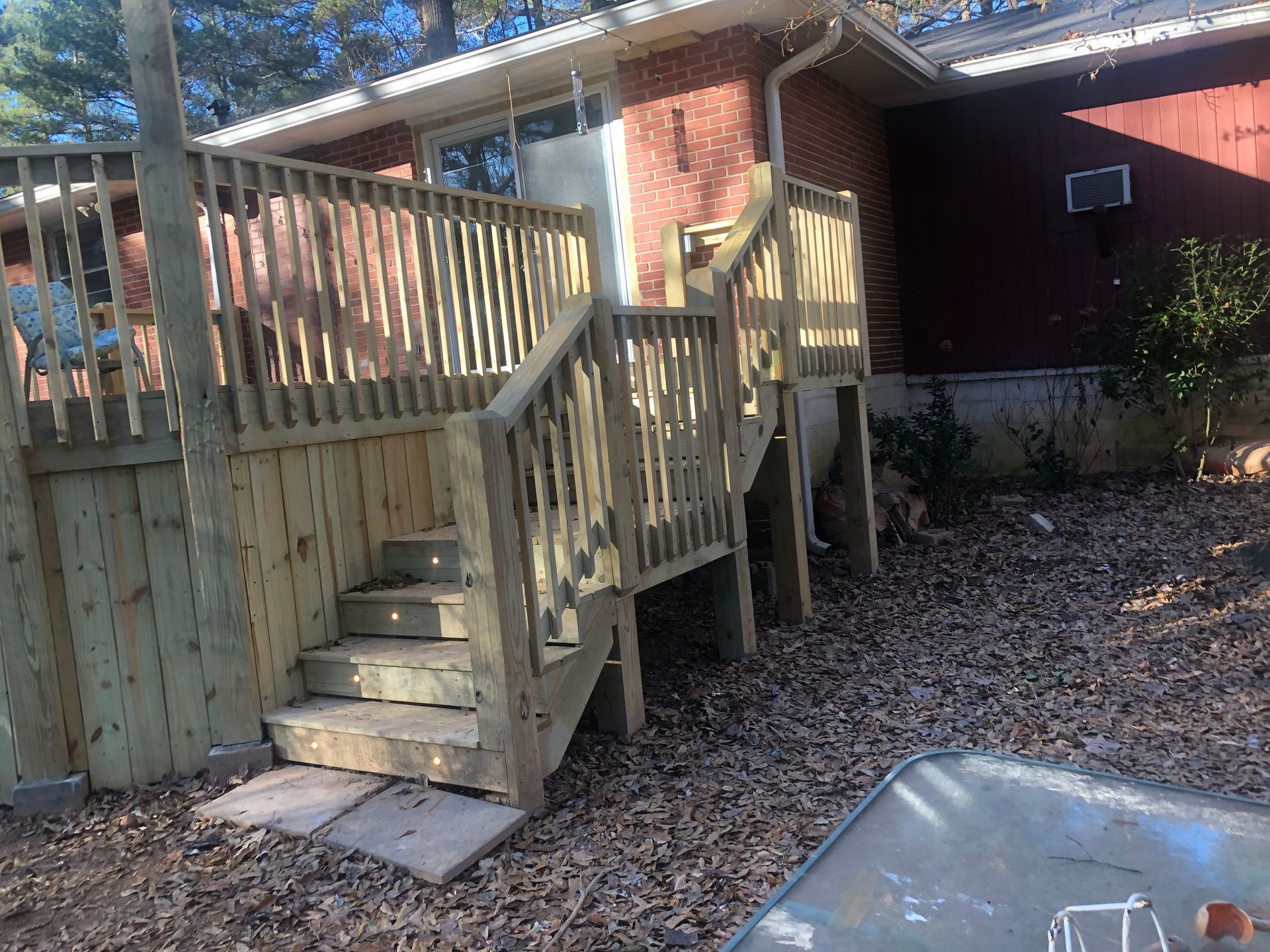 Wooden backyard deck stairs with railing beside a house, beside a paved patio and leaf-covered ground.