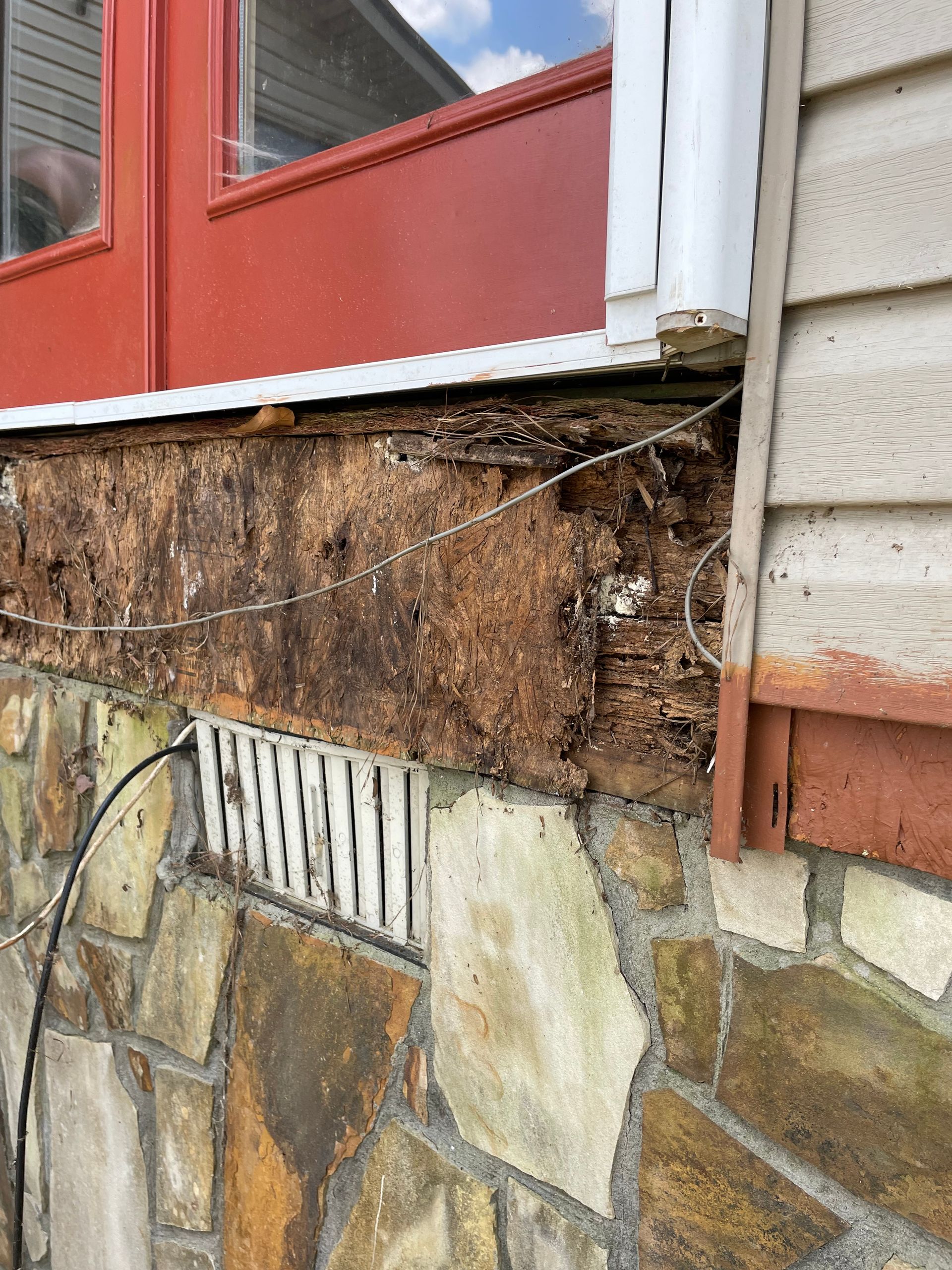 Water-damaged exterior wall with missing siding, exposed wood, and stone veneer beneath a red window.
