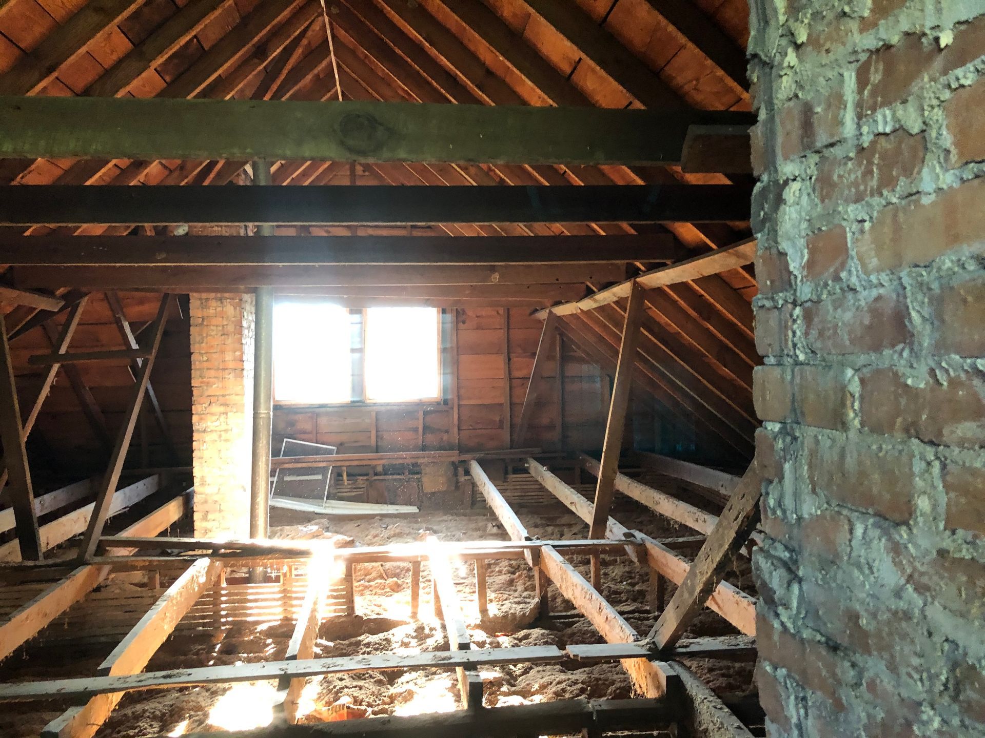Attic interior with exposed wooden rafters, brick chimney, and a bright window at the far end