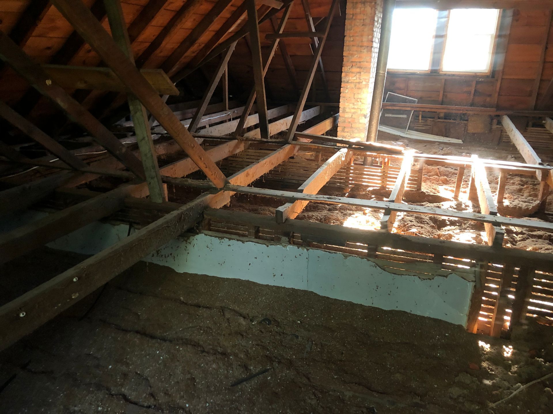 Interior of an attic with exposed wooden beams, sunlight through the roof, and insulation below.