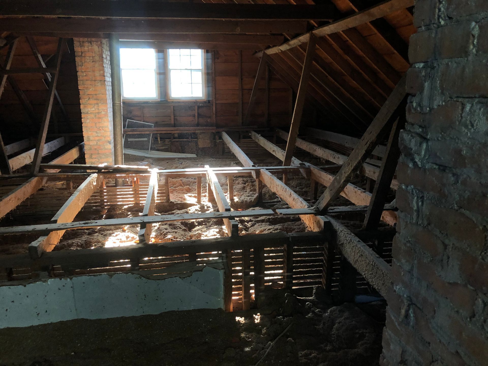 Interior of a damaged attic with exposed rafters, broken floorboards, and sunlight through a small window.