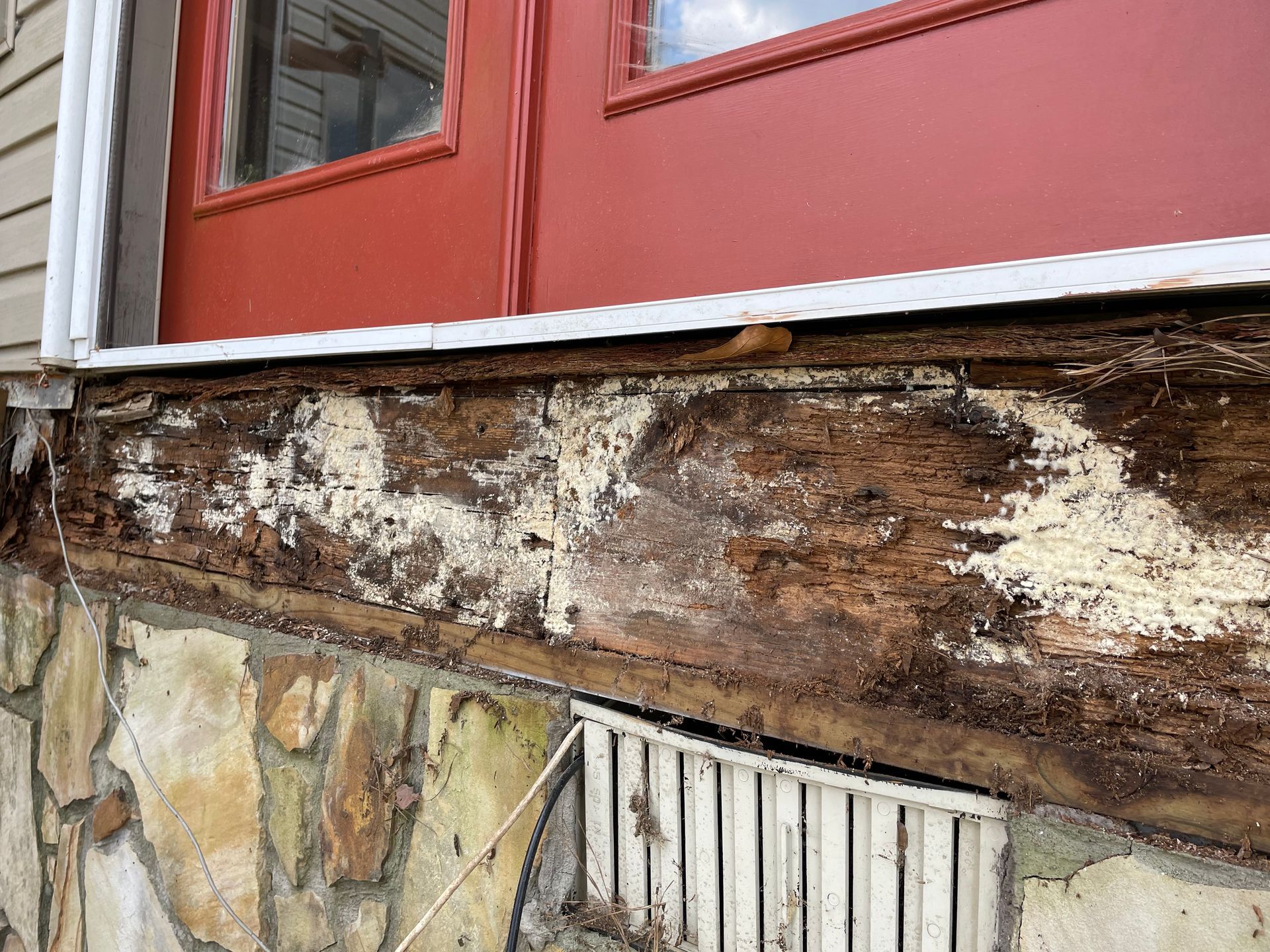Damaged wooden wall beneath a red door with peeling paint and exposed decay