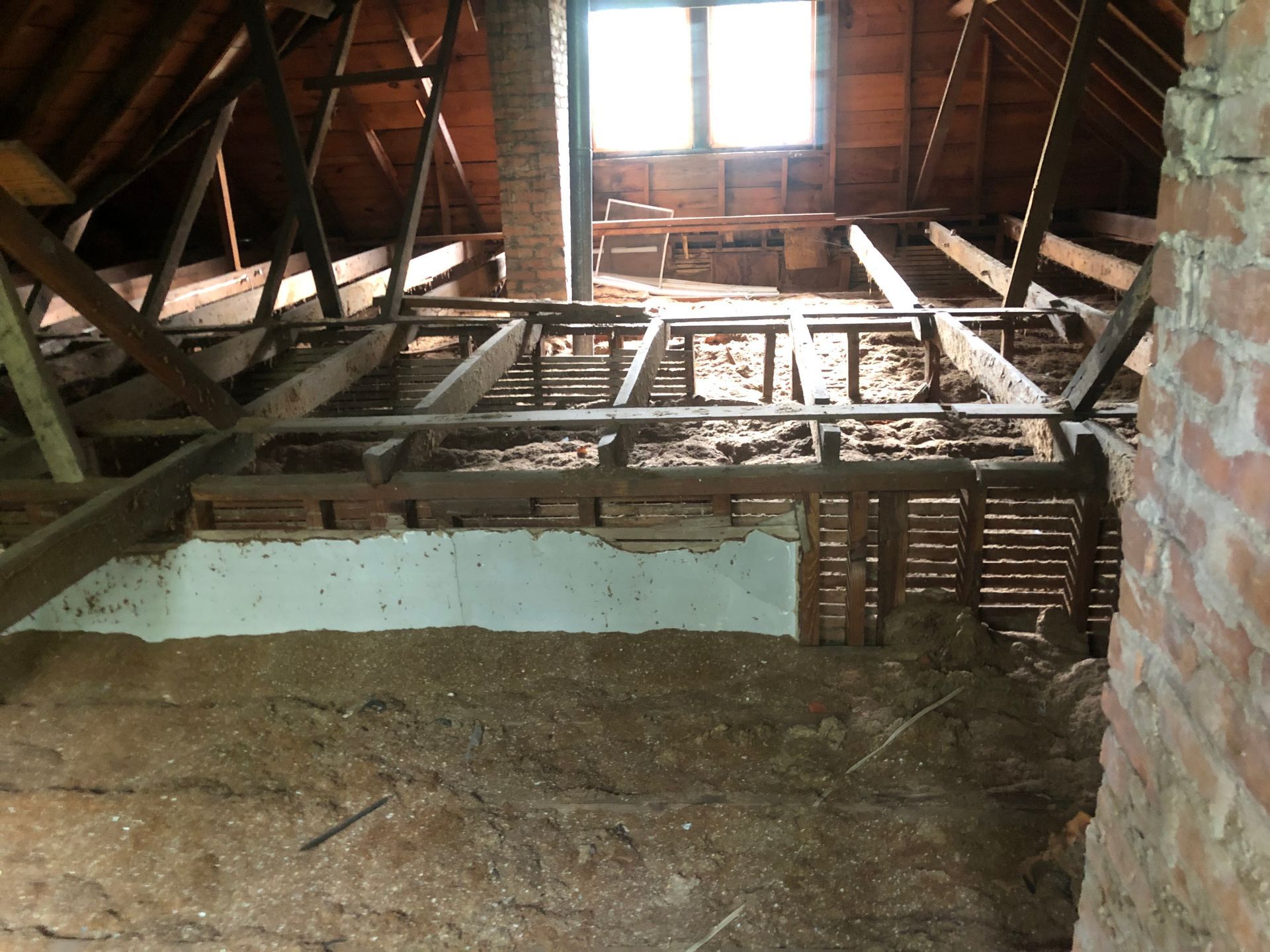 Abandoned attic with exposed wooden beams, a broken floor, and sunlight through a small window.
