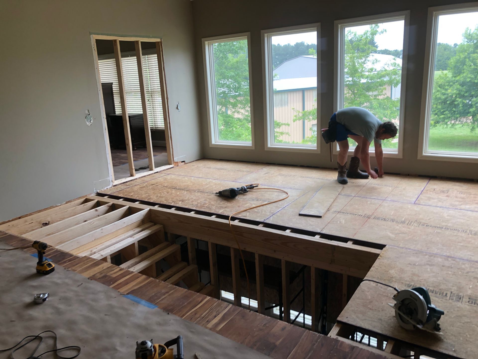 Home renovation with floor joists exposed and a person working on plywood subfloor near large windows