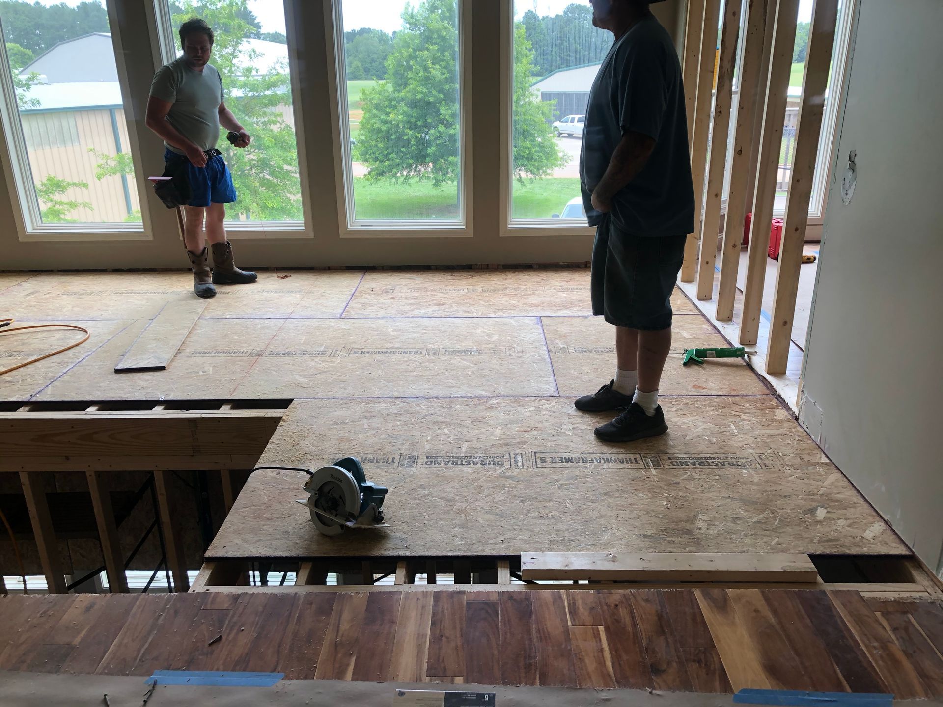 Two people stand on an unfinished floor inside a room with large windows and exposed framing.