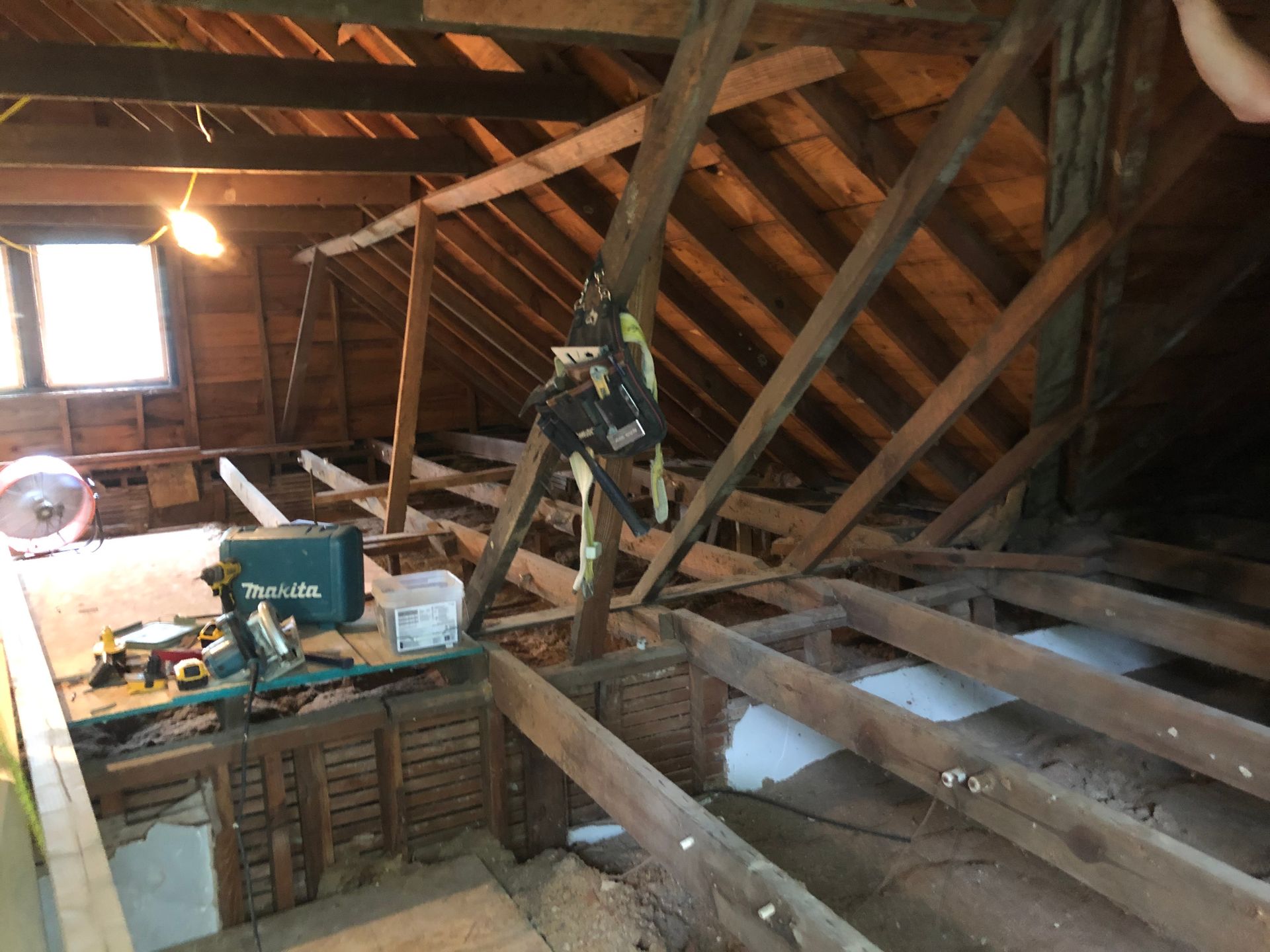 Attic with exposed beams, a circular saw on boards, and a worker on a ladder under a slanted roof.
