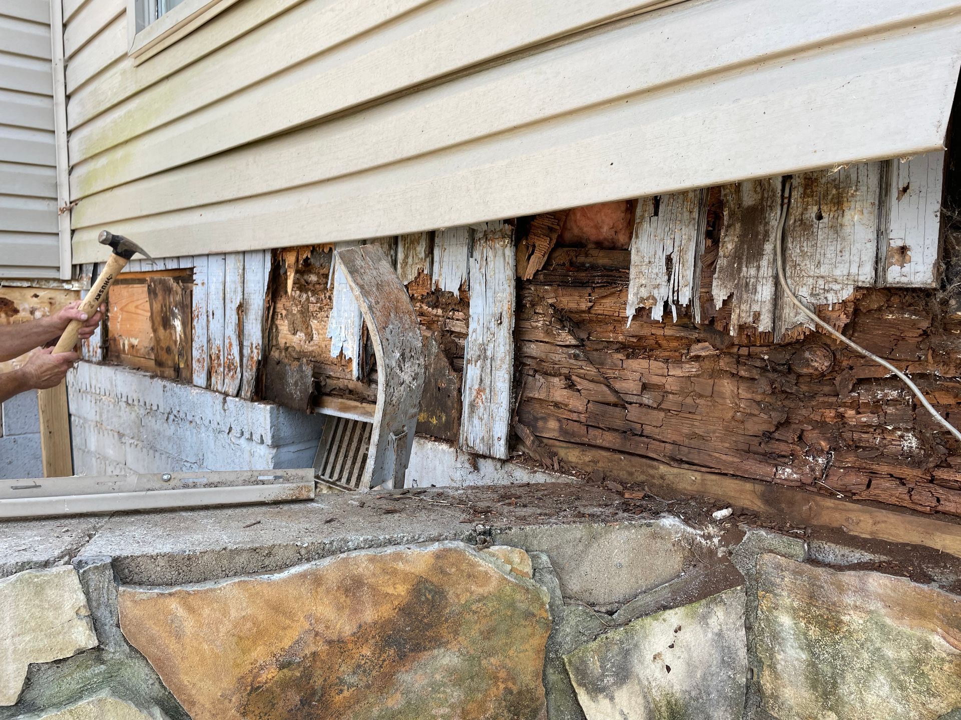 Severely rotted house siding and exposed wood foundation above a stone retaining wall