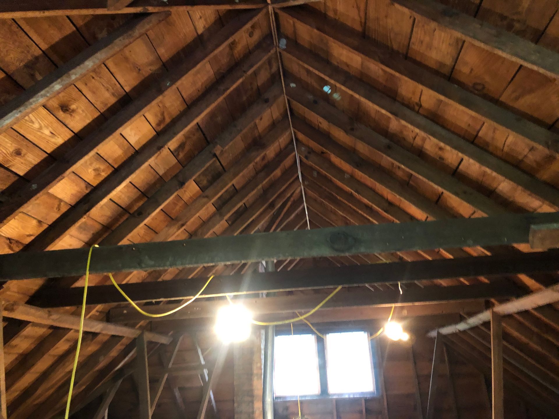 Attic interior with exposed wooden rafters, hanging wires, and a small window with light bulbs below