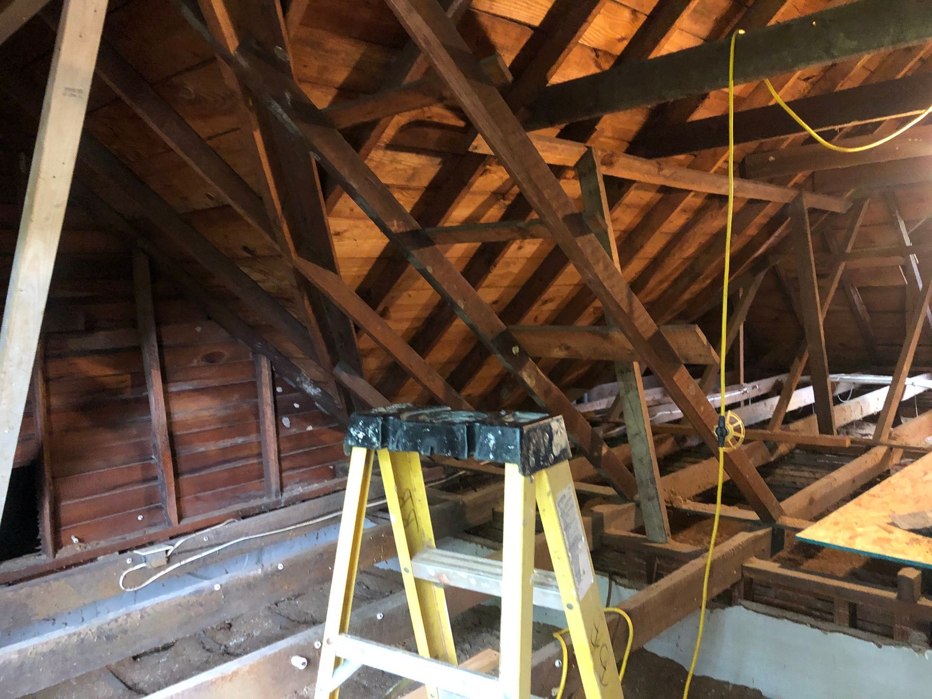 Attic with exposed wooden rafters, a yellow ladder, and hanging wires under construction
