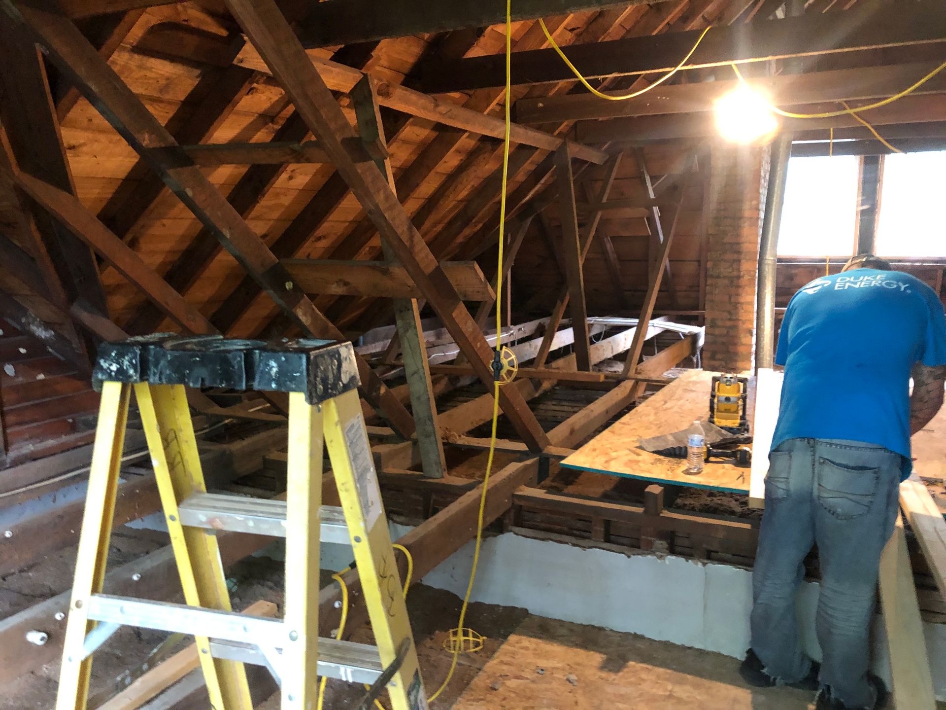 Workers repairing an attic roof with ladders, exposed beams, and a bright work light.