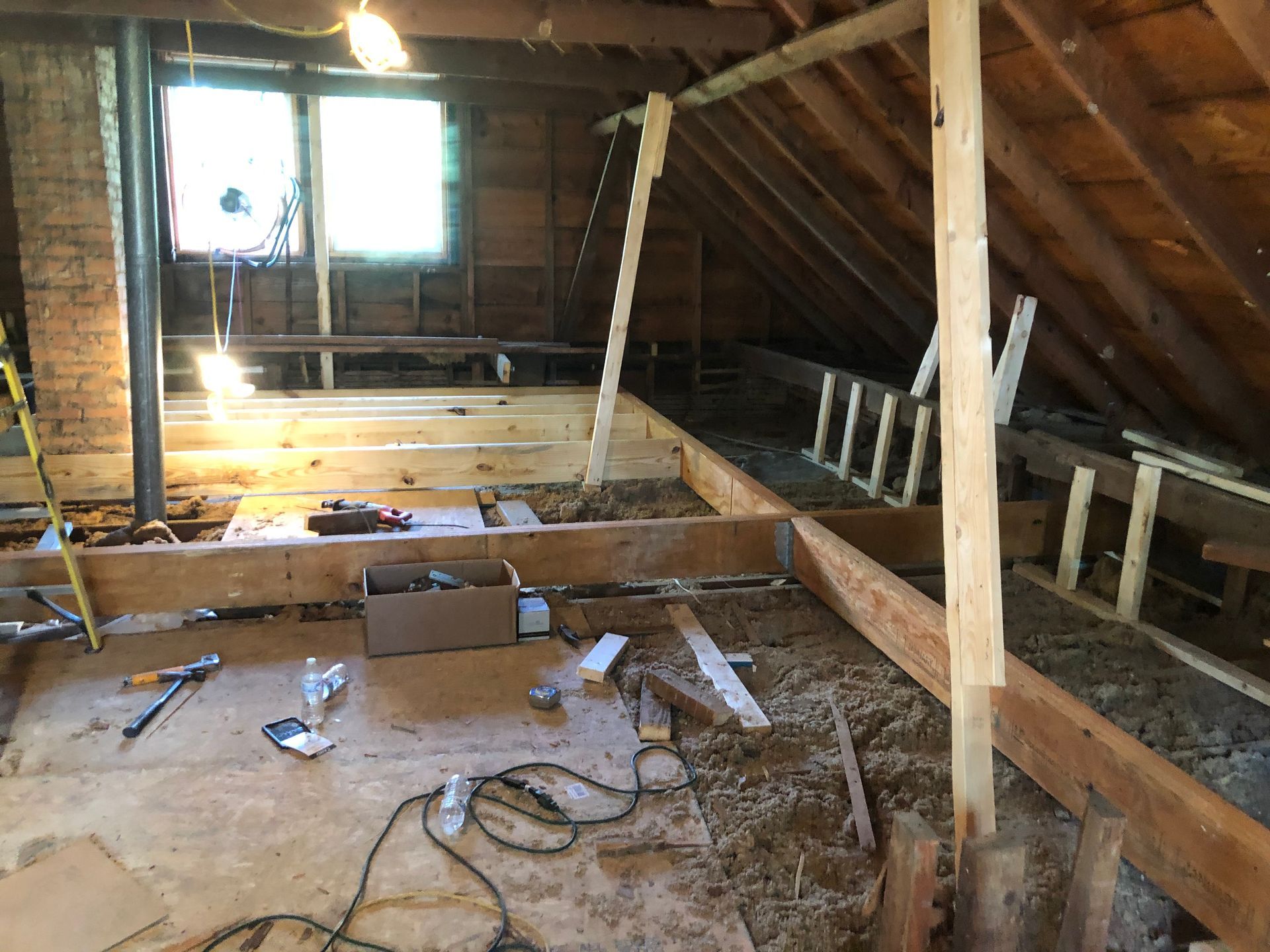 Attic under construction with exposed beams, floor joists, tools, and daylight through a window.