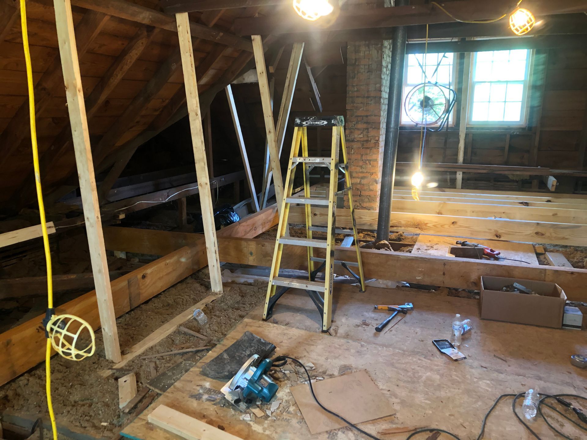 Attic under renovation with ladder, tools, exposed beams, and sunlight through windows