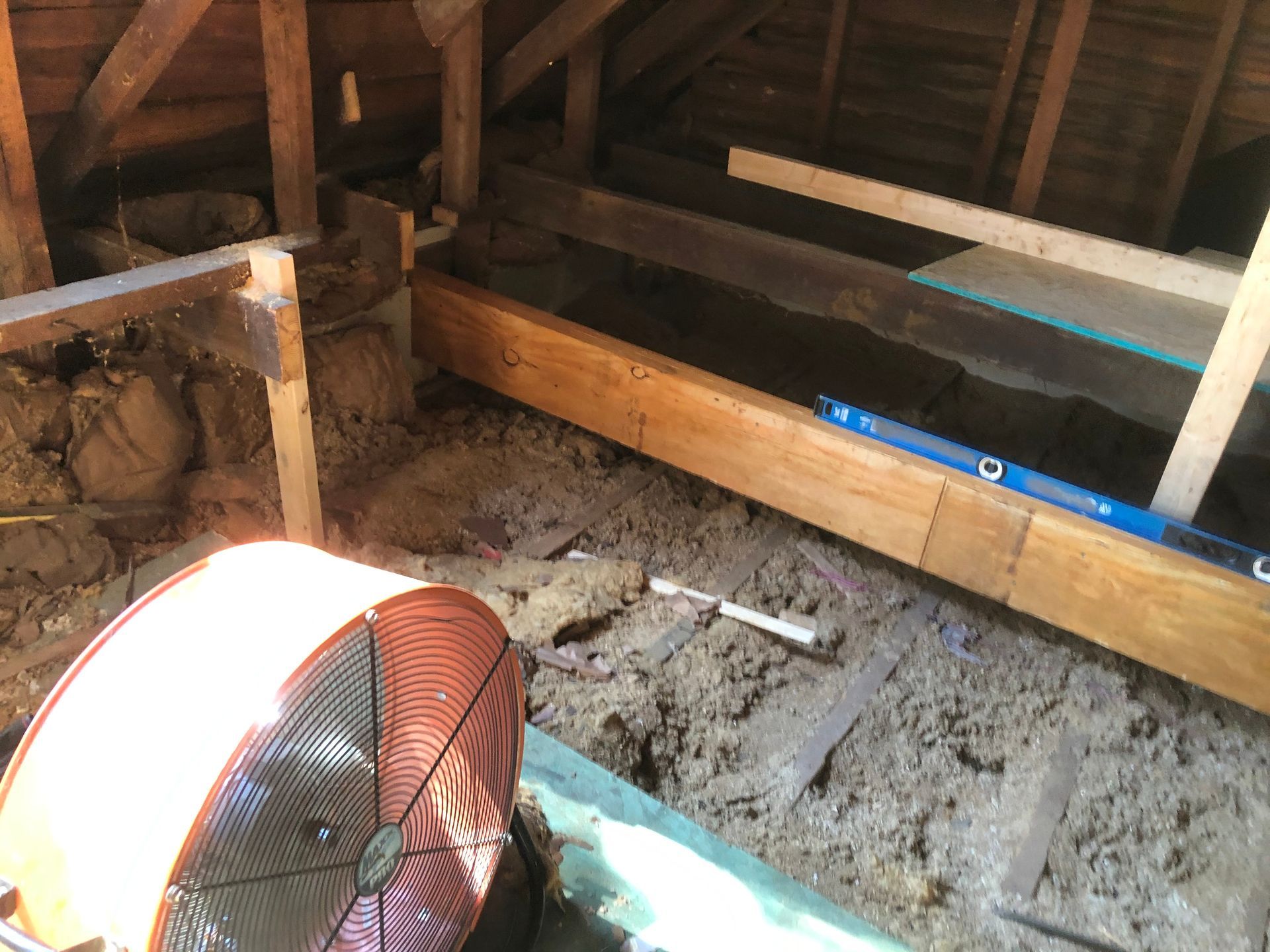 Attic with exposed beams, insulation boards, and a large orange fan on the floor near a dirt-covered area