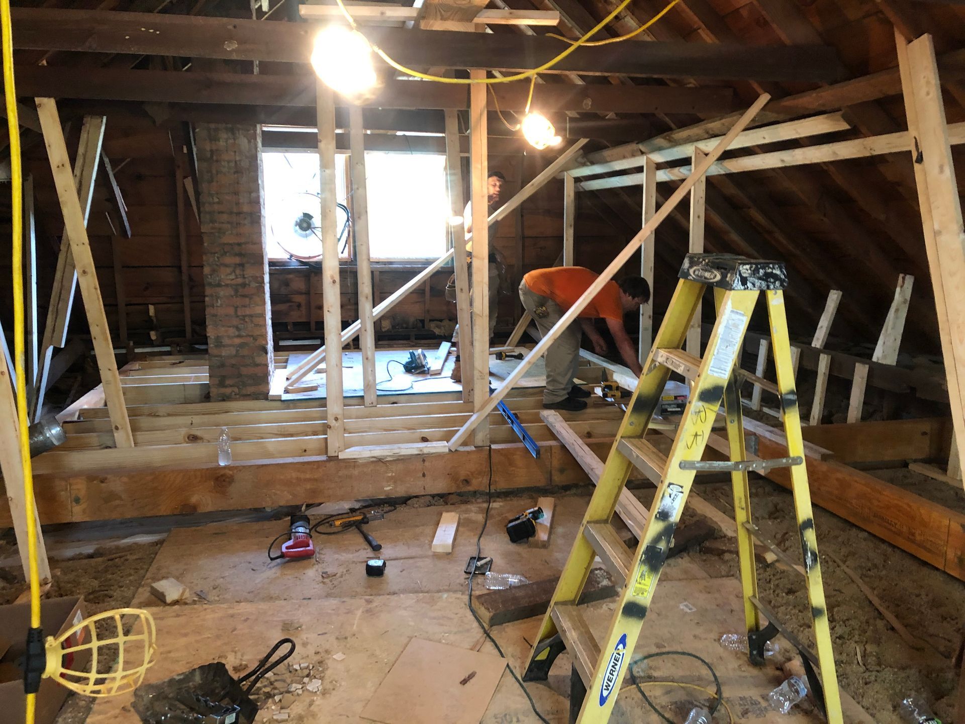 Attic renovation with ladders, tools, exposed beams, and workers under bright lights