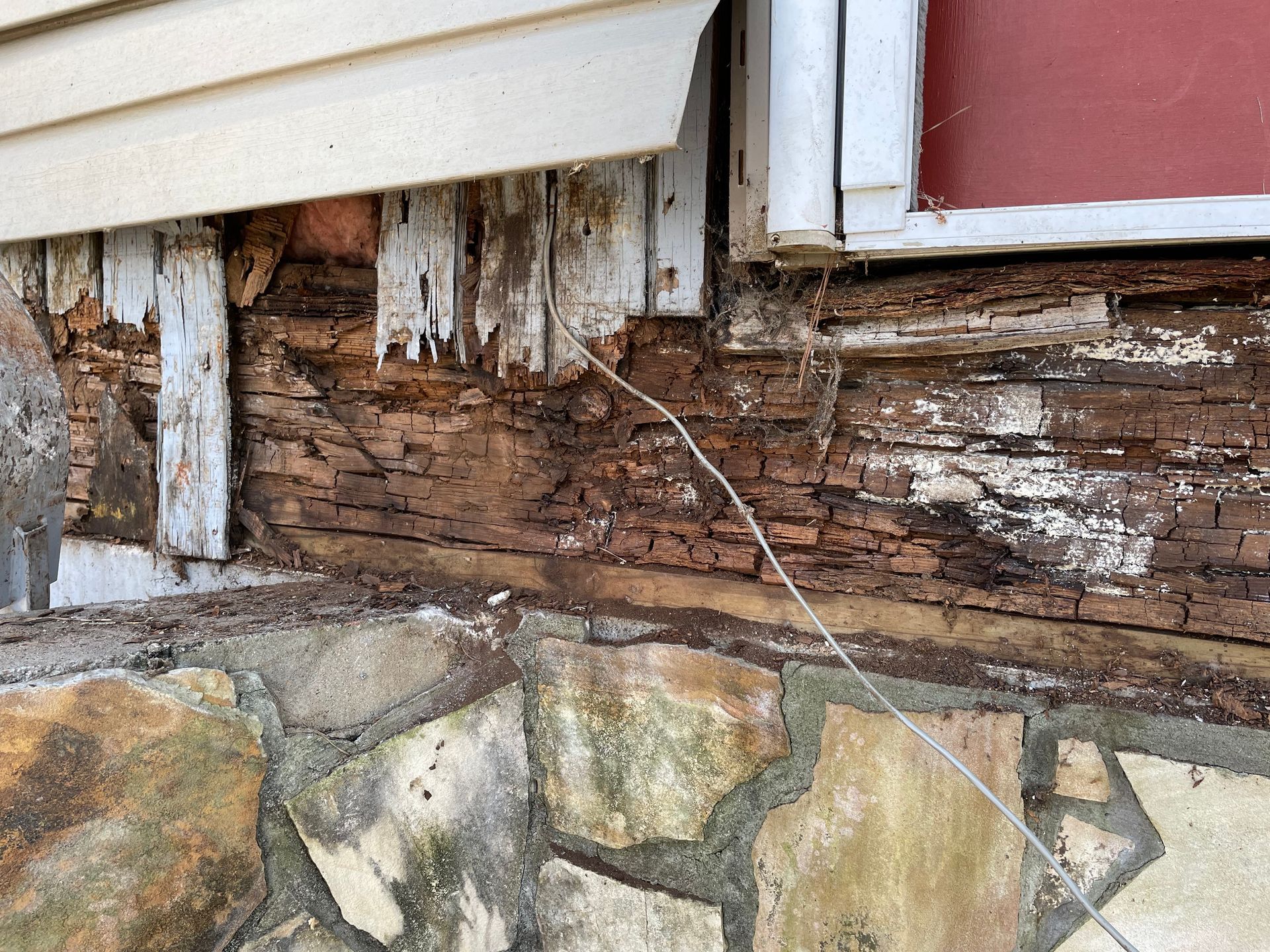 Deteriorated wooden siding and stone foundation with visible rot and peeling paint near a window